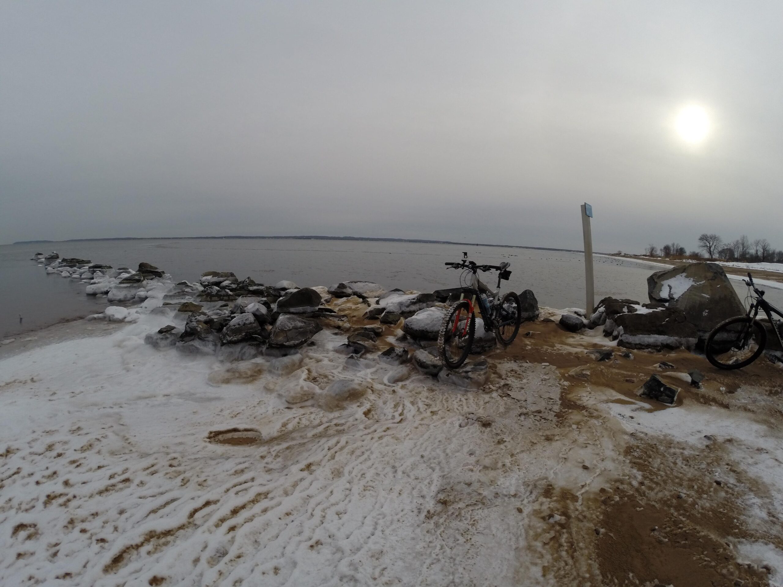 A winter coastal scene featuring two bicycles parked near a snowy shoreline. In the background, a calm body of water extends to the horizon, framed by a rocky outcrop partially covered in snow. The sky is overcast, creating a soft, muted light, with the sun faintly visible. The sandy beach is marked by snow and ice, contributing to the serene winter atmosphere. Staten Island Beach mountain bike trail.