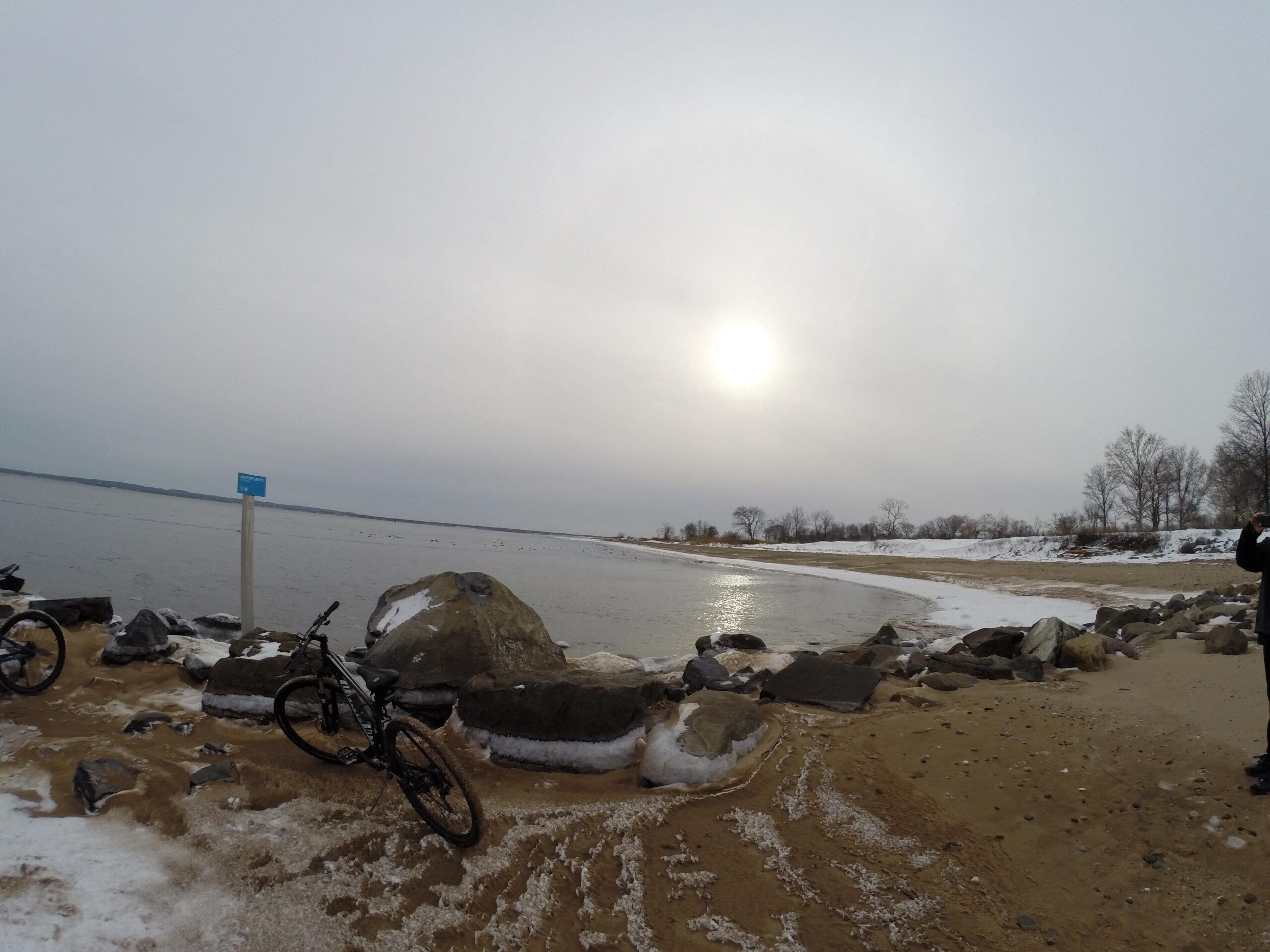 A scenic view of a beach during winter, featuring a calm body of water reflecting a cloudy sky. The shoreline is lined with rocks and patches of snow. In the foreground, a black bicycle is parked on the sandy beach. A blue sign is visible on the left, with trees in the background. The sun is partially obscured by clouds, creating a tranquil atmosphere. Staten Island Beach mountain bike trail.