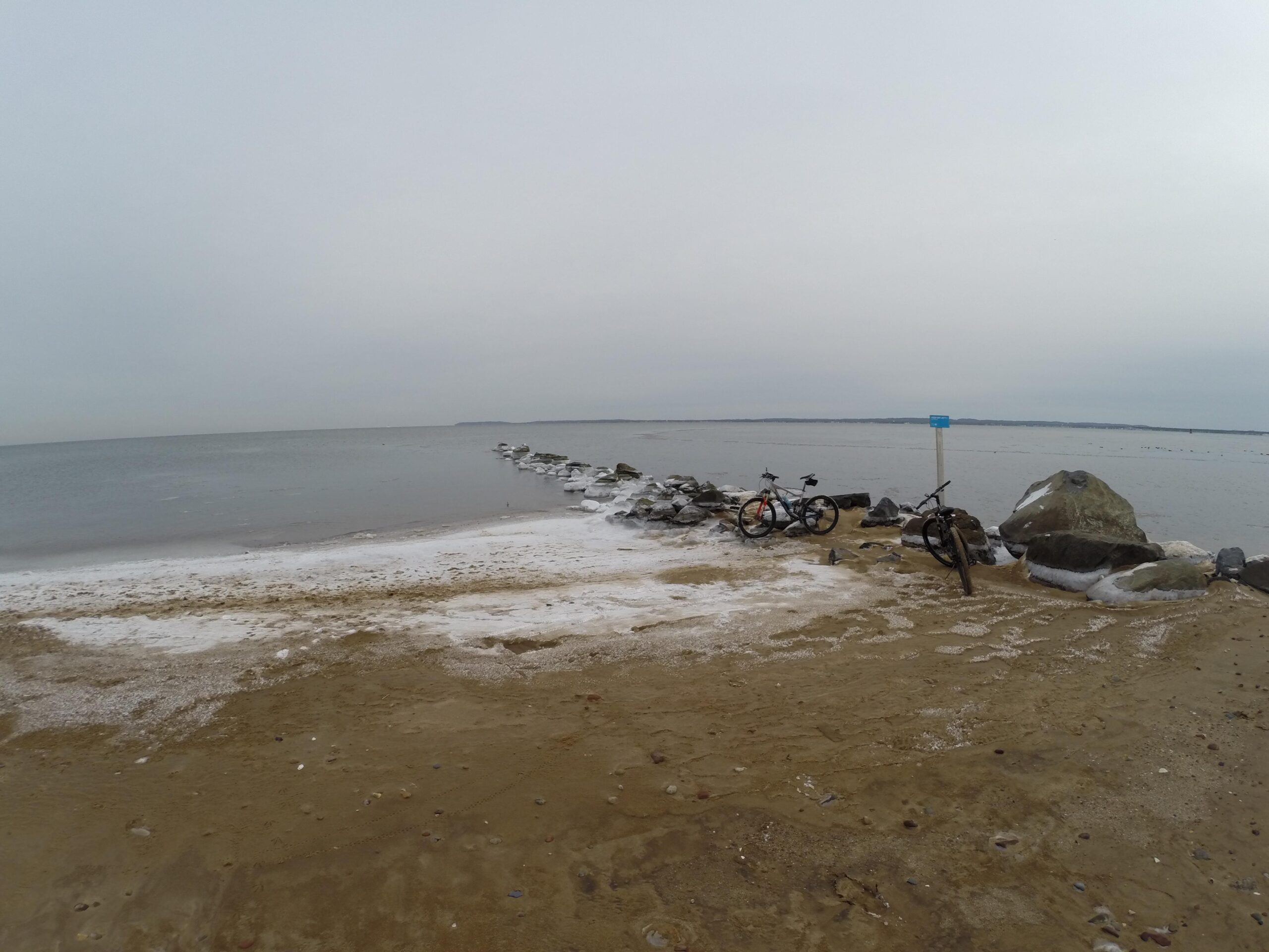 A sandy beach scene with gentle waves lapping at the shoreline, featuring a jetty made of rocks extending into the water. Two bicycles are parked near a large rock on the beach, with a signpost visible in the background. The sky is overcast, creating a calm, muted atmosphere. Staten Island Beach mountain bike trail.