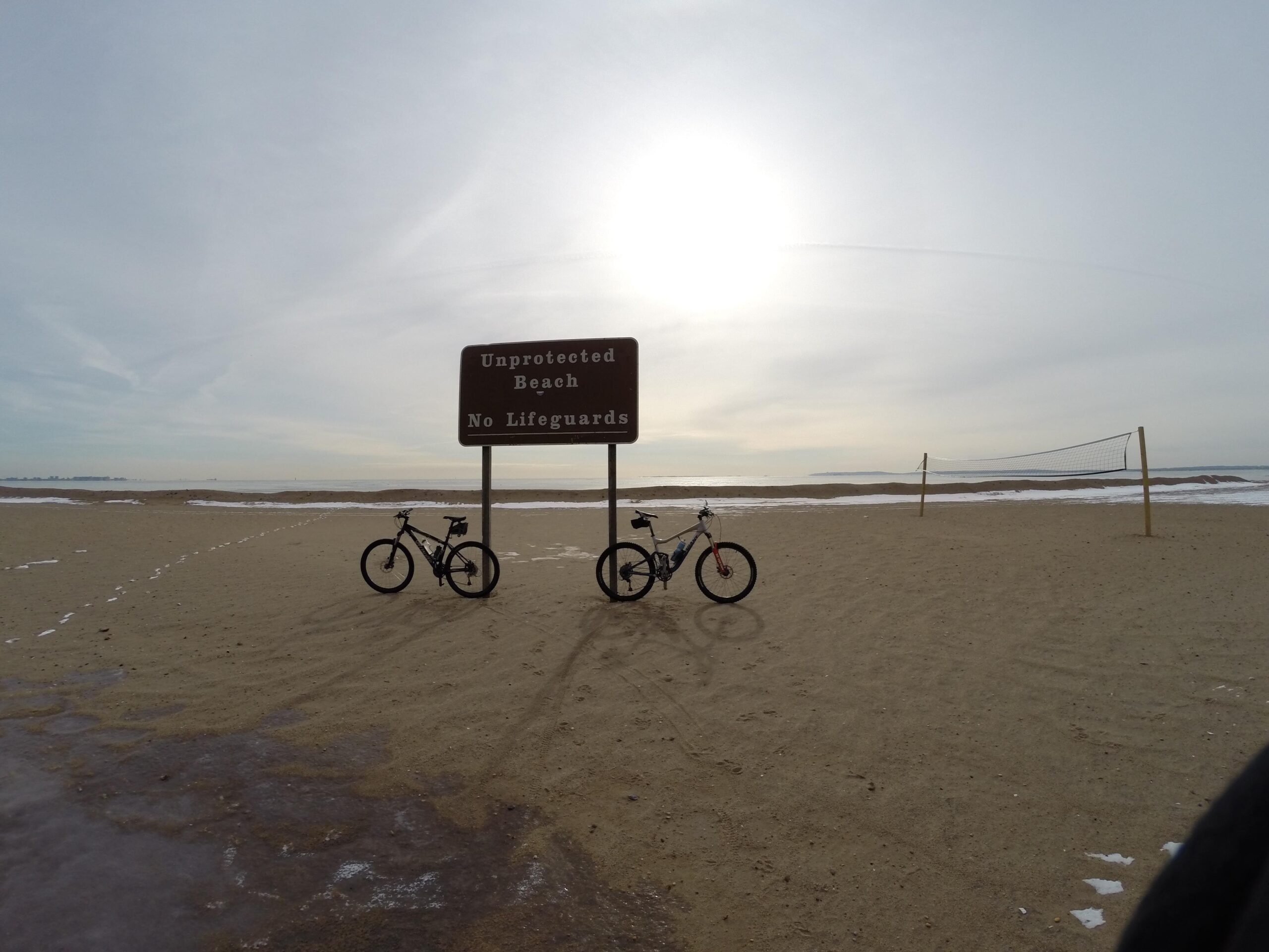 Two bicycles are parked on a sandy beach with a sign that reads "Unprotected Beach, No Lifeguards." In the background, there is a volleyball net, and the sky is partly cloudy with the sun shining brightly. The ocean can be seen in the distance, adding to the serene coastal atmosphere. Gateway Beach mountain bike trail.