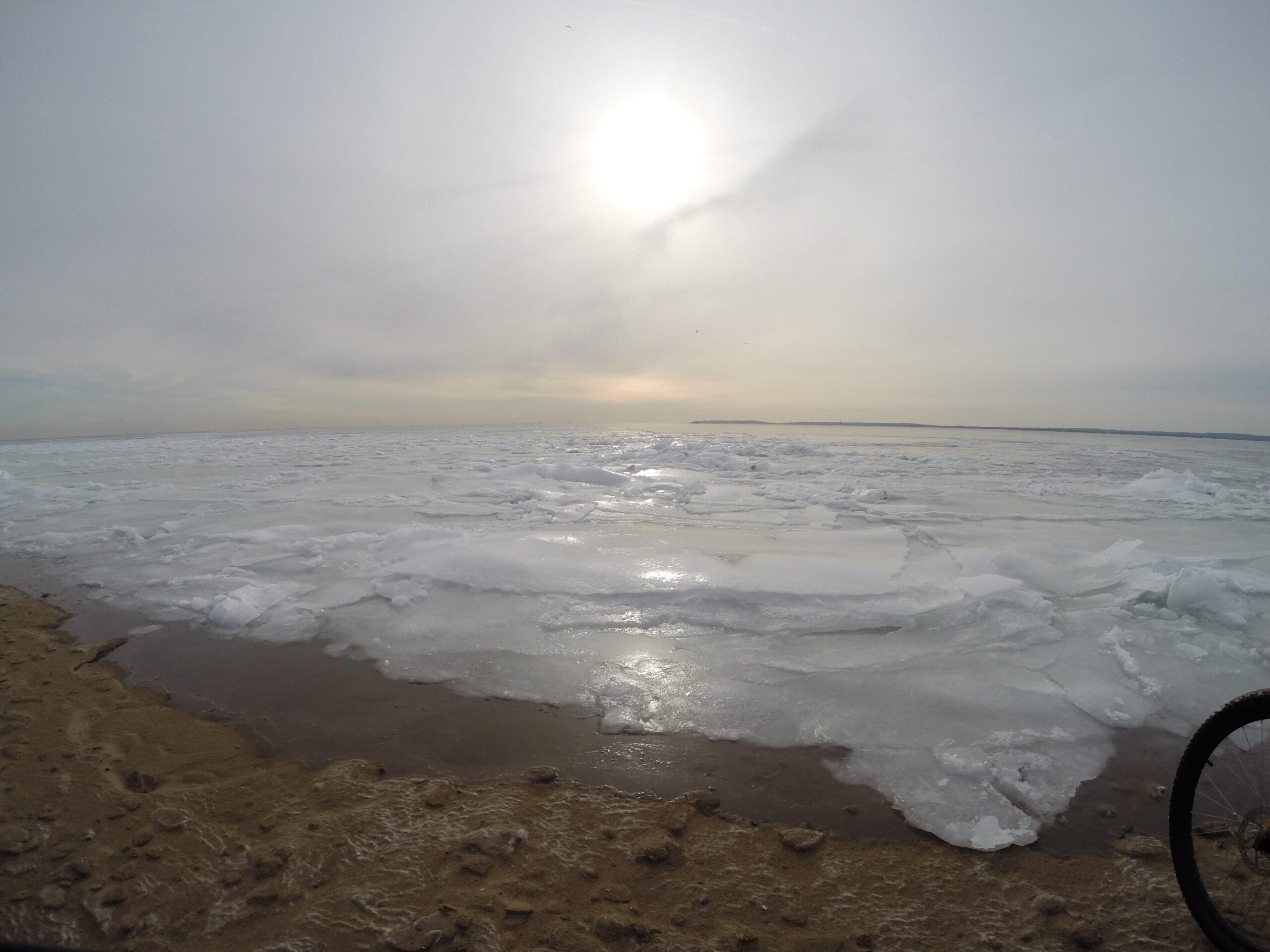 A serene winter landscape featuring a partially frozen body of water, with large chunks of ice floating on the surface. The scene is illuminated by a low sun in a cloudy sky, creating a soft, muted color palette. In the foreground, there is a sandy shoreline with small pebbles visible. The edge of a bicycle is partly visible on the right side of the image. Gateway Beach mountain bike trail.