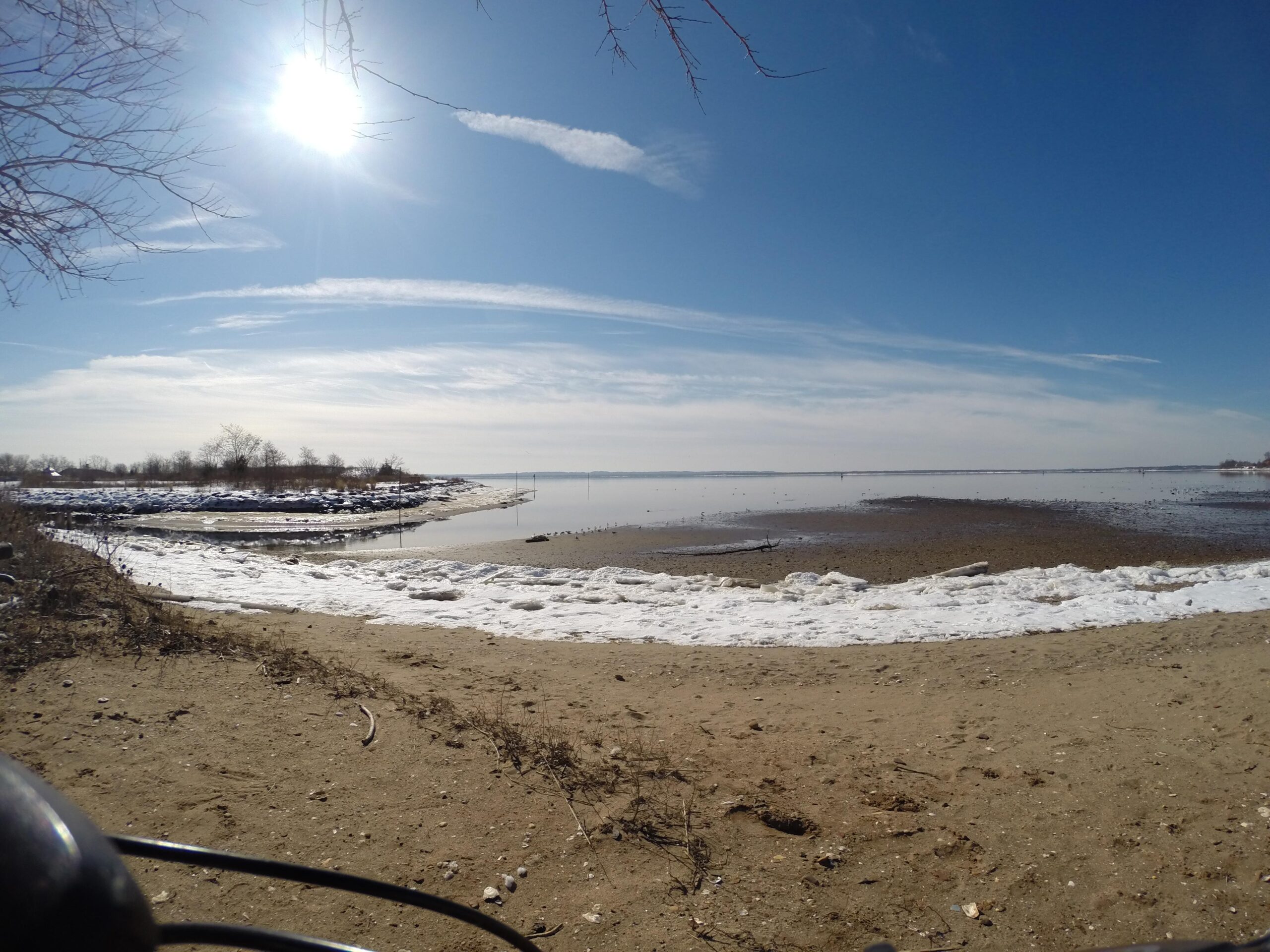 A picturesque shoreline view on a sunny day, featuring a mix of sandy beach and patches of snow. The calm water reflects the clear blue sky with wispy clouds, while the sun shines brightly overhead. In the background, sparse trees line the bank, creating a serene atmosphere. Beach Long Pond Area mountain bike trail.