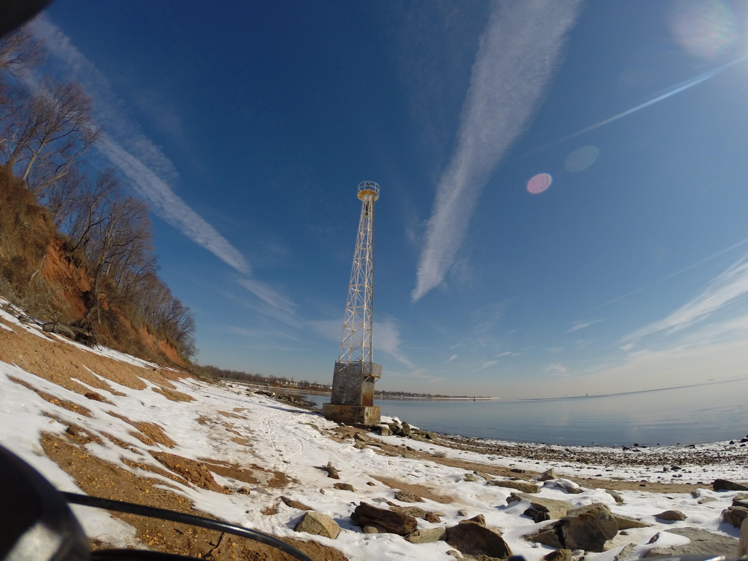 Towering water tower against a blue sky with wispy clouds, situated along a snowy beach beside calm waters, with rocky shoreline and trees in the background. Beach Long Pond Area mountain bike trail.