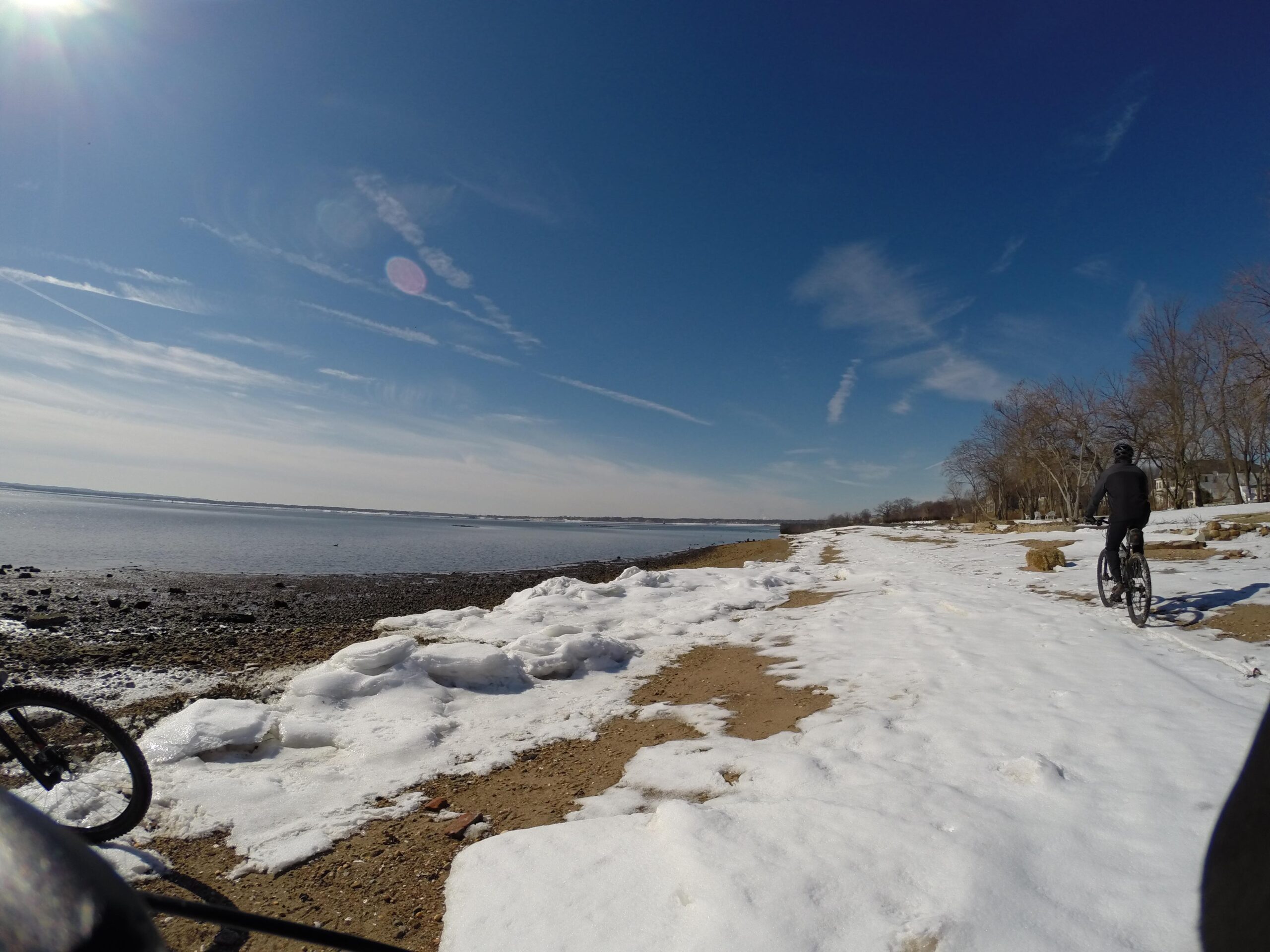 A scenic view of a snowy beach with a cyclist riding along the shoreline. The foreground features patches of snow and rocky terrain, while the calm water stretches into the distance under a clear blue sky streaked with clouds. Trees line the edge of the beach, contributing to the serene winter landscape. Beach Long Pond Area mountain bike trail.