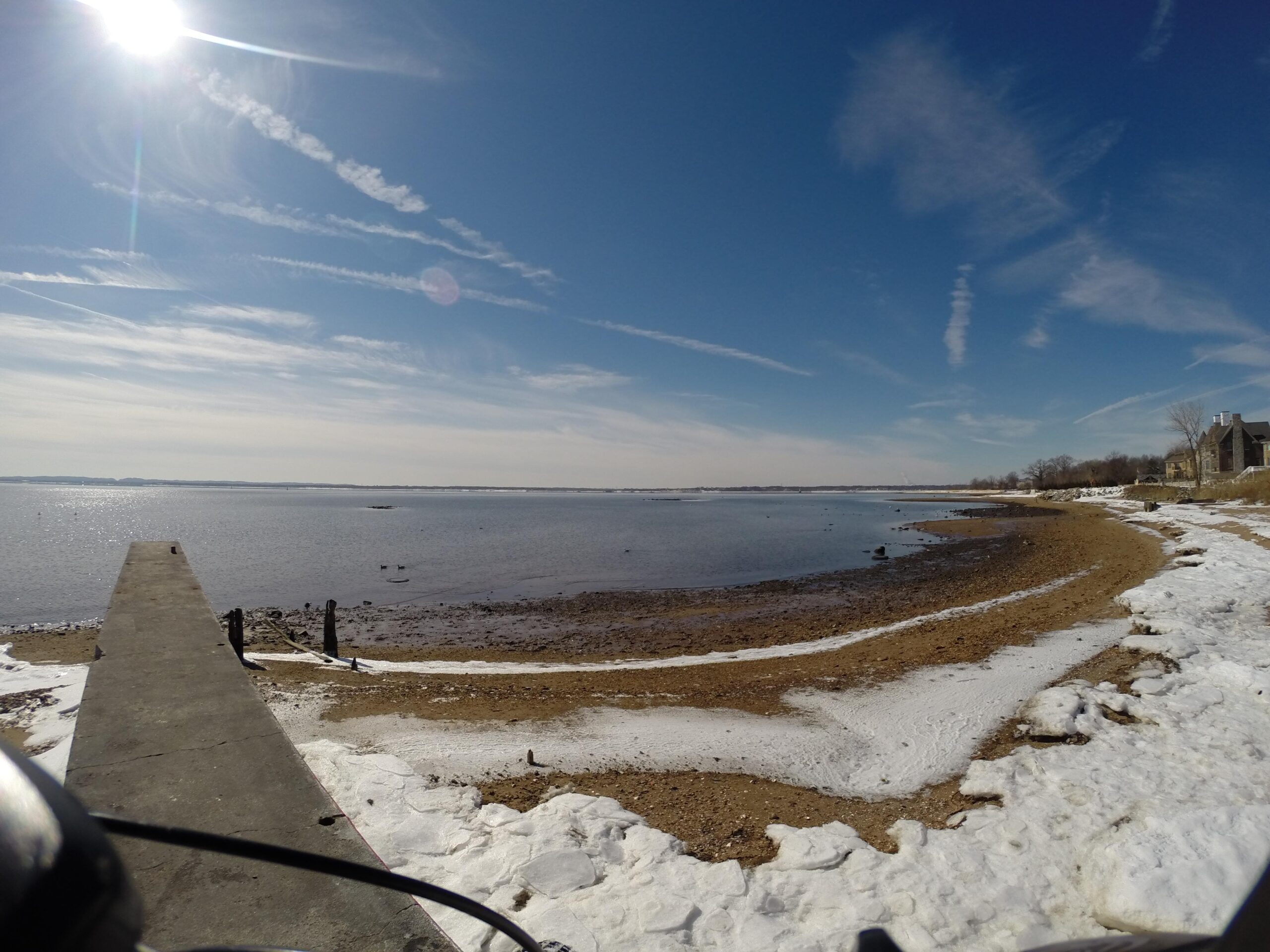 A scenic view of a winter shoreline, showing a calm body of water reflecting the blue sky. The sun shines brightly overhead, casting light on the sandy beach mixed with patches of snow. A concrete pier extends into the water, and the shoreline is lined with snow and gravel. Wispy clouds stretch across the sky, creating a serene atmosphere. Beach Long Pond Area mountain bike trail.