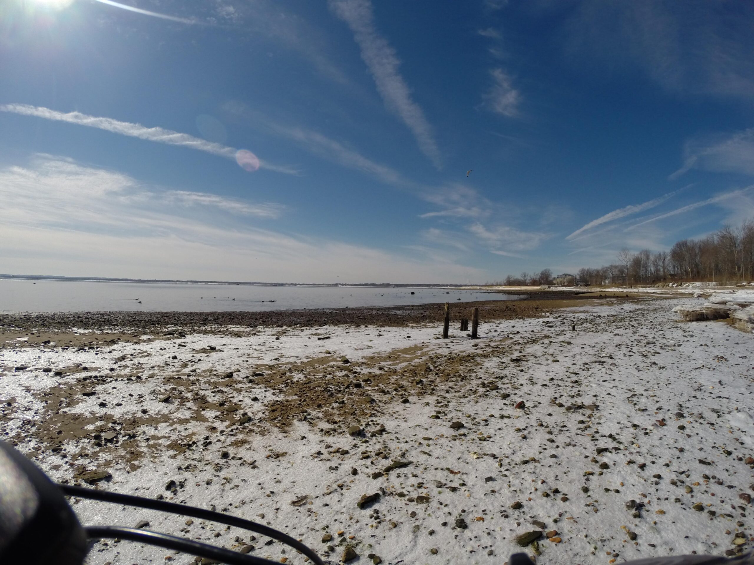 A coastal view showing a rocky shoreline under a clear blue sky with wispy clouds. In the foreground, there are pebbles and small rocks scattered on the beach, while gentle waves can be seen in the distance. A few birds are visible on the water, and the scene captures a tranquil, natural environment. Beach Long Pond Area mountain bike trail.