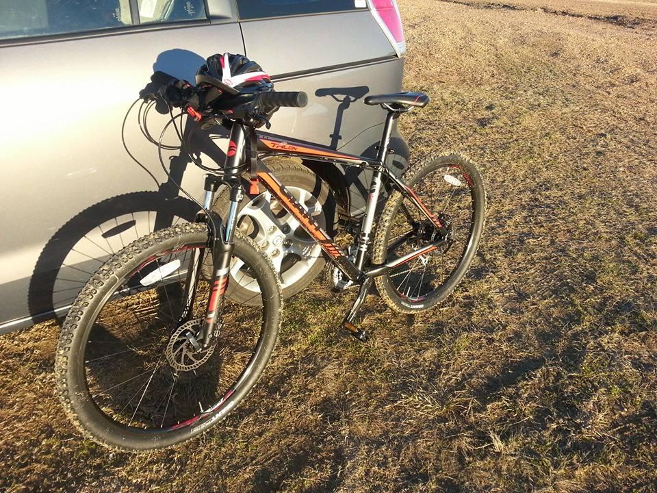 Giant Talon 27.5: A black and orange mountain bike leaning against a silver car, parked on a grassy area. The bike has thick tires and a bike helmet is resting on the handlebars, with the sun casting long shadows.