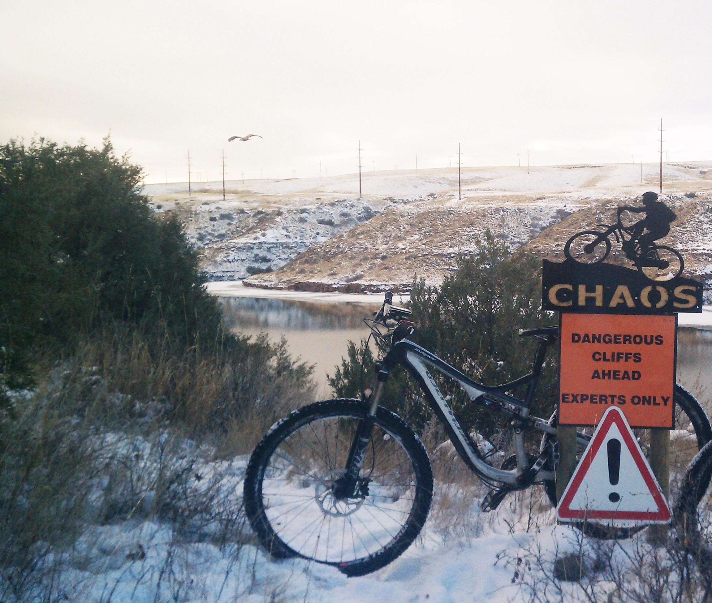Specialized Stumpjumper FSR Comp 29: A mountain bike parked next to a warning sign that reads "CHAOS - DANGEROUS CLIFFS AHEAD - EXPERTS ONLY." The background features a snowy landscape with a river and barren hills, and a bird is flying in the sky.