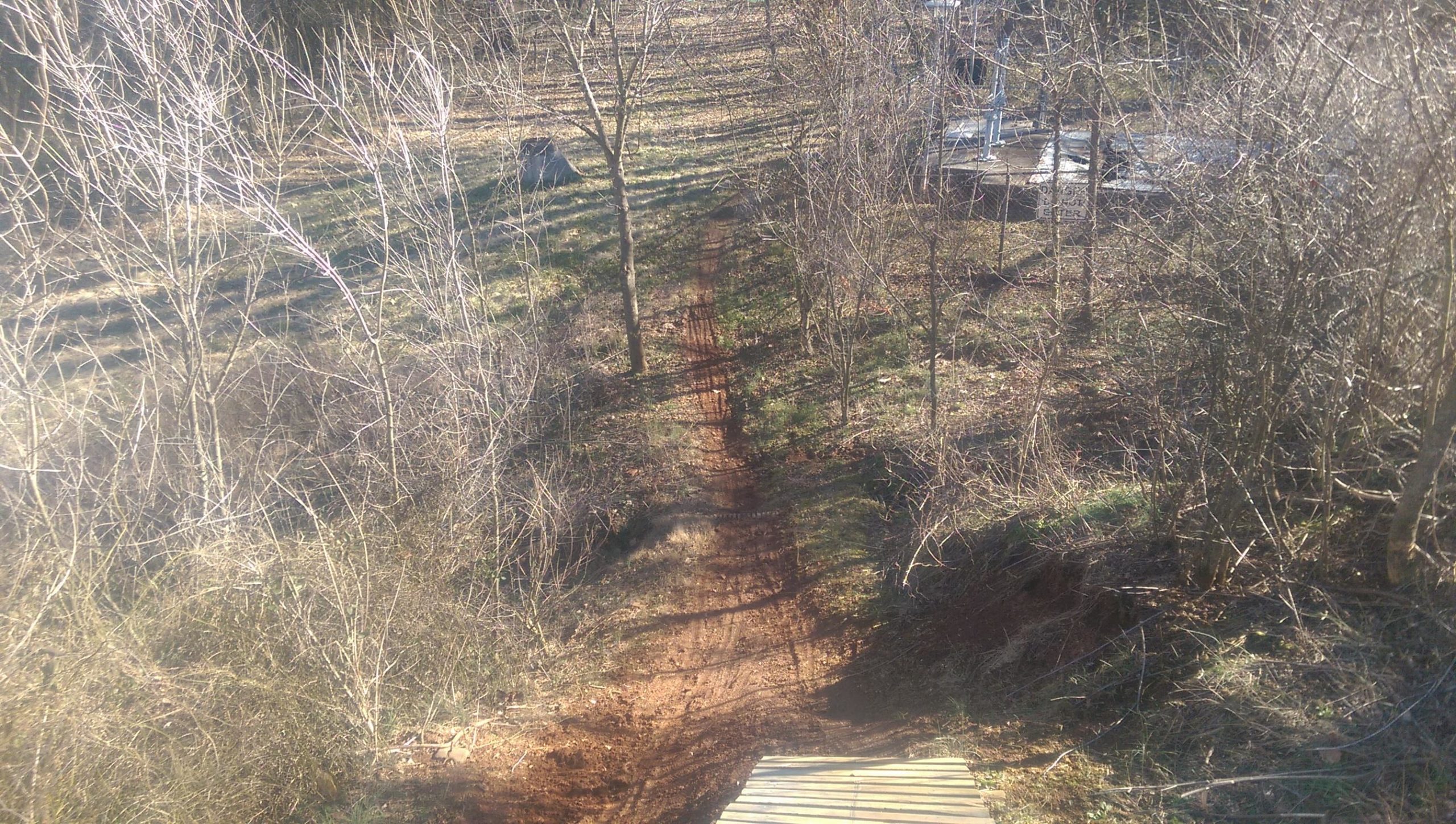 A winding dirt trail runs through a forested area, with sparse trees and underbrush lining the path. The ground shows signs of use, and a wooden bridge or platform is visible at the bottom of the image. Sunlight casts shadows on the trail, suggesting a cool, midday environment. Signal Hill mountain bike trail.