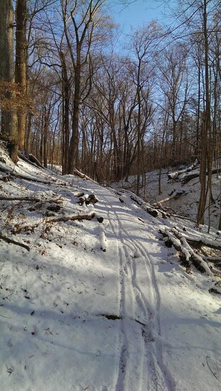 A snow-covered trail winding through a forest, surrounded by bare trees and fallen branches. The bright blue sky is visible above, creating a serene winter landscape. Patapsco Valley State Park (Avalon Area) mountain bike trail.