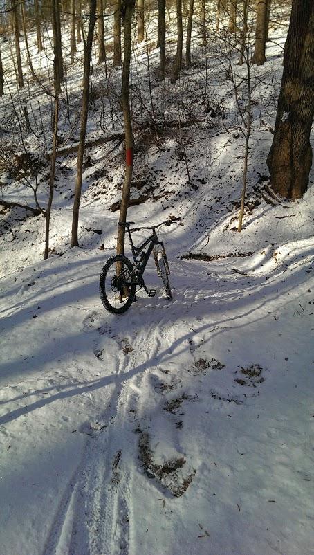 A mountain bike parked on a snow-covered trail in a wooded area. The scene includes trees with bare branches and a slight incline, showing tire tracks and footprints in the snow. Patapsco Valley State Park (Avalon Area) mountain bike trail.