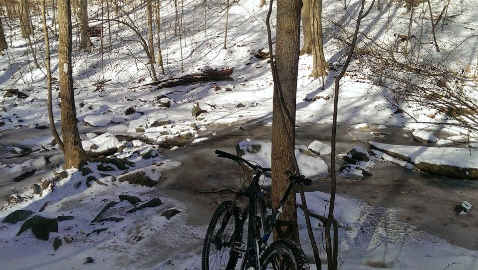Two mountain bikes are leaning against a tree in a snowy forest setting. In the background, a partially frozen creek flows over rocks, surrounded by snow-covered ground and bare trees. Soft sunlight filters through the trees, creating a serene winter landscape. Patapsco Valley State Park (Avalon Area) mountain bike trail.