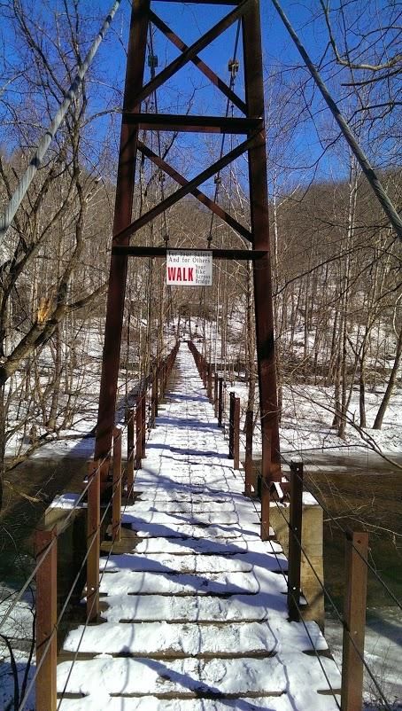 A snow-covered suspension bridge stretches across a river, with bare trees lining the banks. A sign attached to the bridge reads "WALK." The scene is set against a clear blue sky. Patapsco Valley State Park (Avalon Area) mountain bike trail.