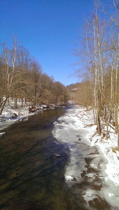 A serene winter landscape featuring a gently flowing river bordered by bare trees. Snow and ice partially cover the riverbanks, while a clear blue sky fills the background. Patapsco Valley State Park (Avalon Area) mountain bike trail.