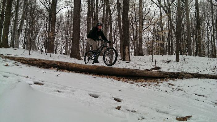 A person riding a mountain bike over a fallen log in a snowy forest. The surrounding area is covered in a layer of snow, and tall trees are visible in the background. Patapsco Valley State Park (Avalon Area) mountain bike trail.