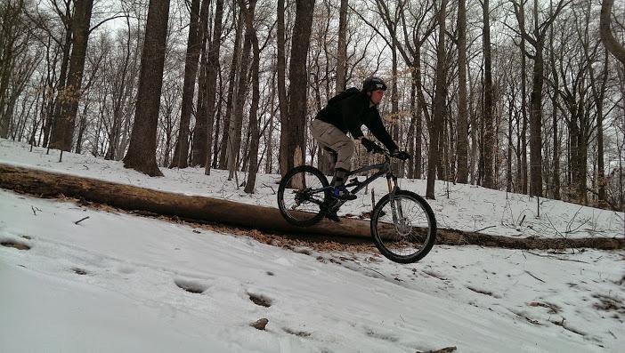 A mountain biker navigating over a fallen log in a snowy forest. The rider, dressed in winter attire, is mid-jump with snow-covered ground and bare trees surrounding the scene. Patapsco Valley State Park (Avalon Area) mountain bike trail.
