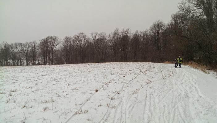 A snow-covered field with tire tracks leading through it, bordered by a line of bare trees under a cloudy sky. A person wearing a bright yellow jacket is seen riding a fat bike along the edge of the field. Patapsco Valley State Park (Avalon Area) mountain bike trail.