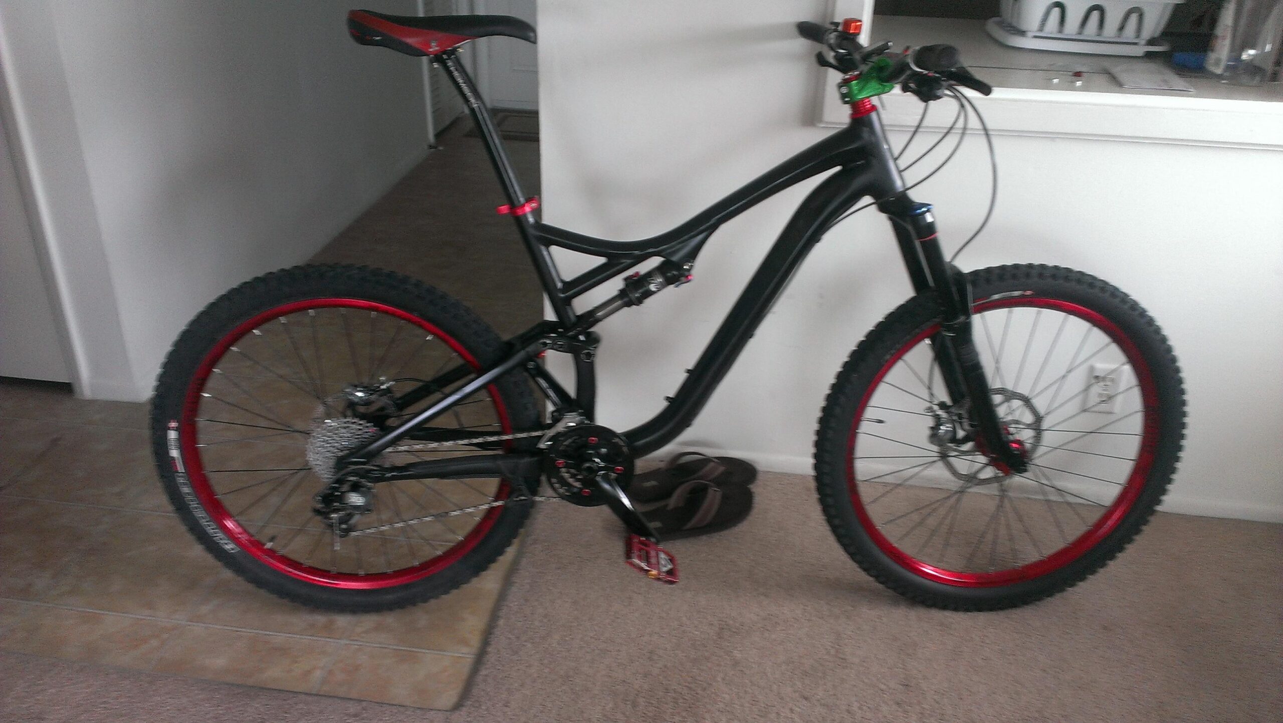 Specialized Stumpjumper FSR Comp EVO: A black mountain bike with red accents, featuring thick tires, a front suspension fork, and a modern frame design, positioned on carpet indoors, next to a pair of flip-flops.