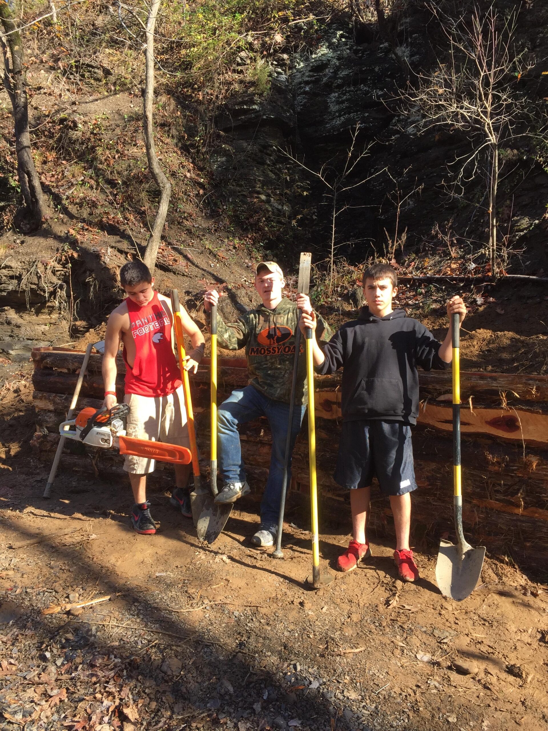 Three young men pose outdoors with various tools, including a chainsaw and shovels. They are standing in front of a pile of logs in a forested area, with trees and rocky terrain in the background. The scene appears to depict a day of physical work or conservation efforts. Spadra Creek Nature Trail mountain bike trail.