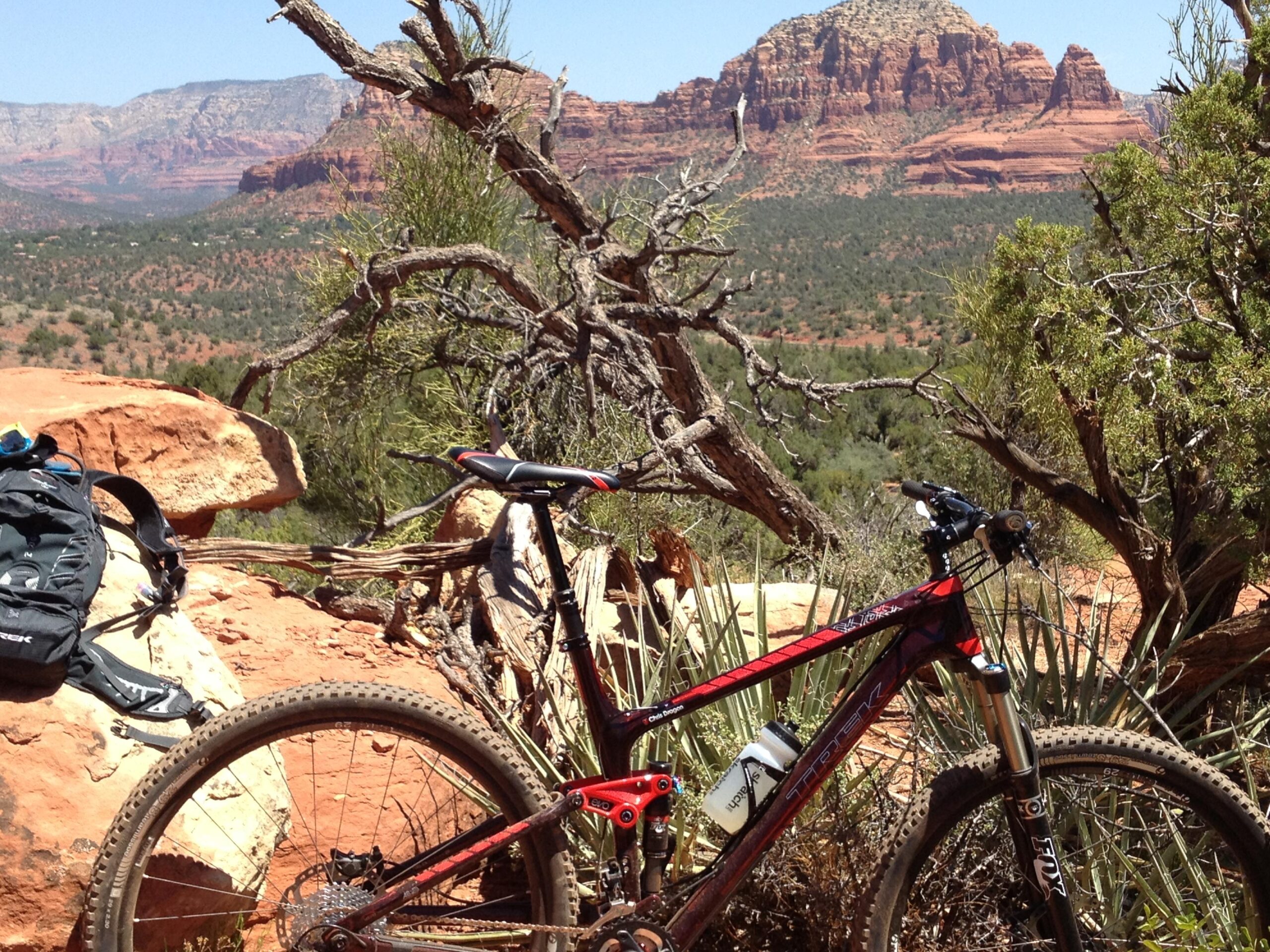 Trek Fuel EX 9.9 29 XX1: A mountain bike parked on a rocky ledge with a scenic view of red rock formations and a green valley in the background. A backpack is positioned nearby, and desert vegetation surrounds the area under a clear blue sky.