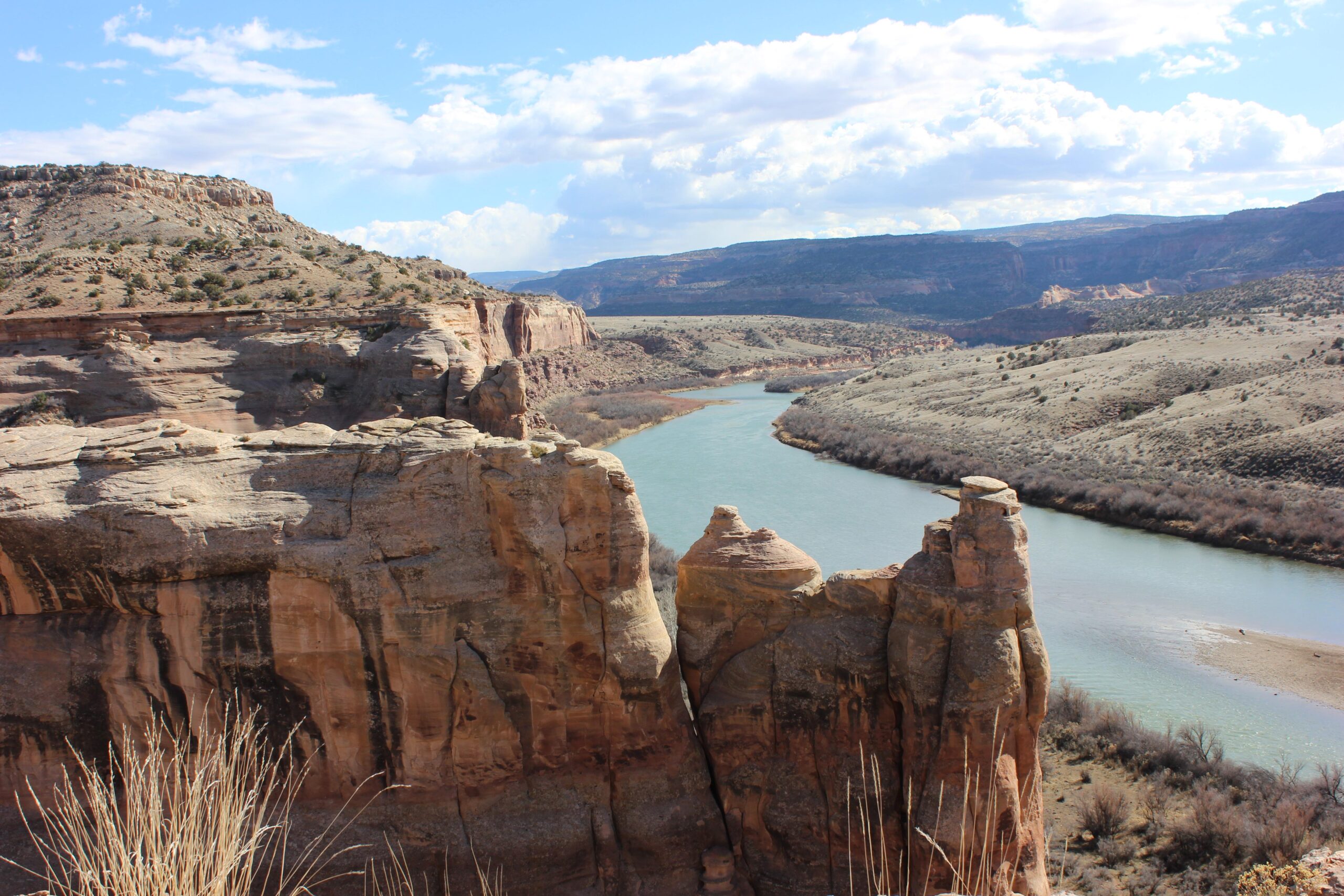 A panoramic view of a river winding through a rugged canyon landscape, featuring dramatic rock formations and a sparse, grassy foreground. The sky is partly cloudy, with sunlight illuminating the rocky terrain and highlighting the contours of the hills in the background. Kokopelli Trail mountain bike trail.