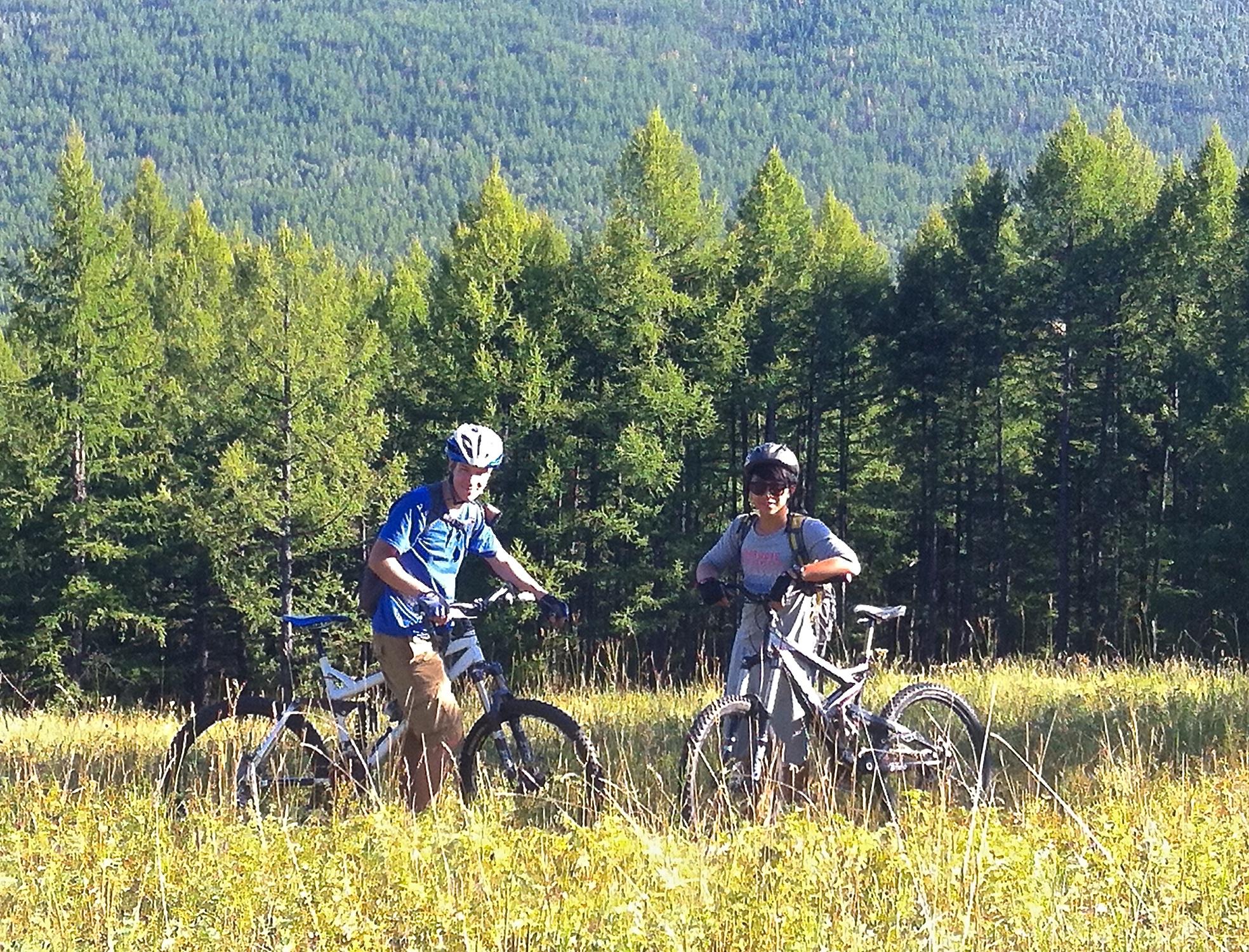 Two mountain bikers pose for a photo in a grassy area surrounded by tall trees and mountains in the background. One biker, wearing a blue shirt and helmet, stands beside a white bicycle, while the other, dressed in a gray shirt with a backpack and also wearing a helmet, stands next to a gray bicycle. The scene is bright and sunny, showcasing a peaceful outdoor setting. Bulgan Range North mountain bike trail.