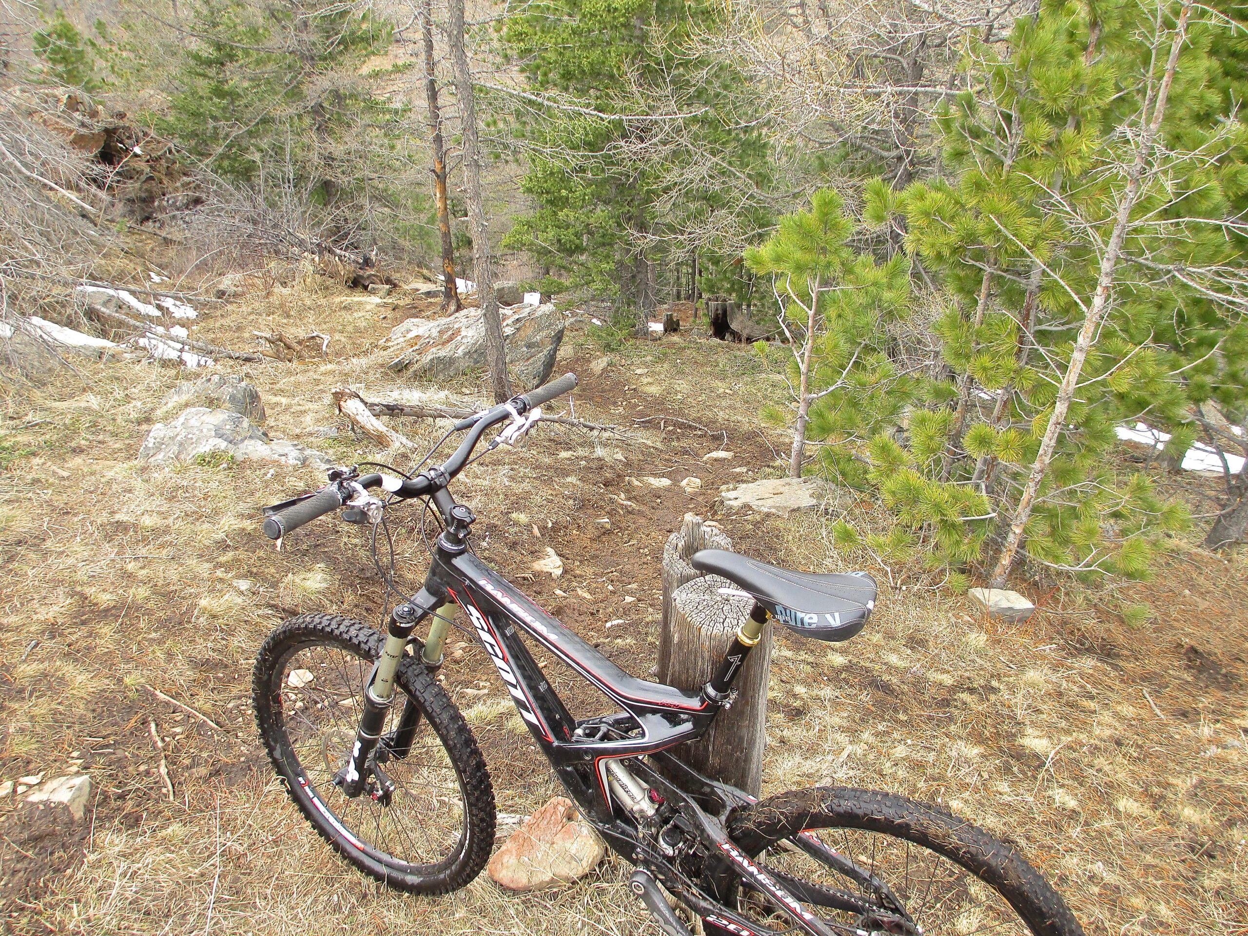 A mountain bike is parked next to a wooden post on a dirt trail surrounded by sparse vegetation and scattered rocks, with trees in the background. The ground is mostly dry with some patches of grass, indicating an outdoor setting suitable for biking. Nukht to Zaisan mountain bike trail.
