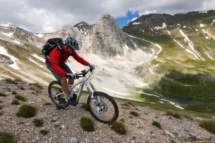 A mountain biker navigating a rocky uphill trail in a mountainous landscape, wearing a helmet and a red long-sleeve shirt, with green hills and snow-capped peaks in the background.