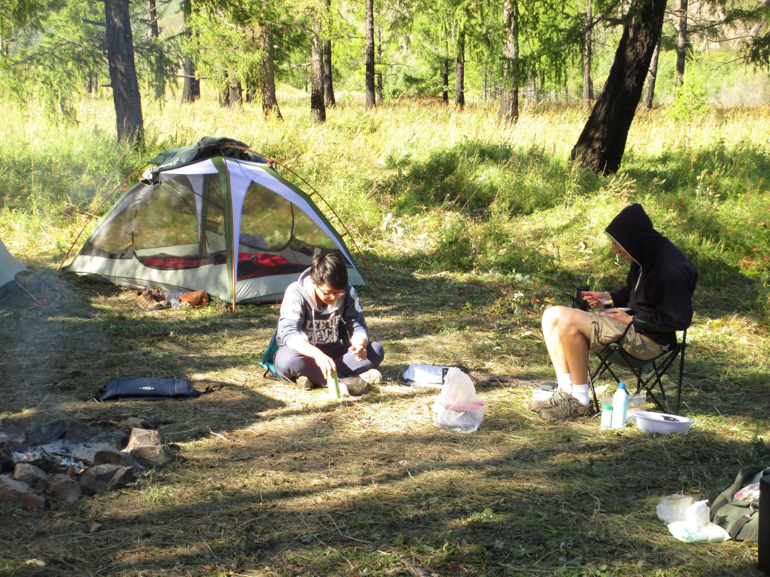 Two individuals are sitting at a campsite in a wooded area. One person is crouched and preparing food, while the other is seated in a camping chair, engaged with a mobile device. A tent is set up nearby, and a fire pit with some remnants of a fire is visible on the ground. The surrounding tall grass and trees suggest a serene outdoor environment. Khargana Gol mountain bike trail.