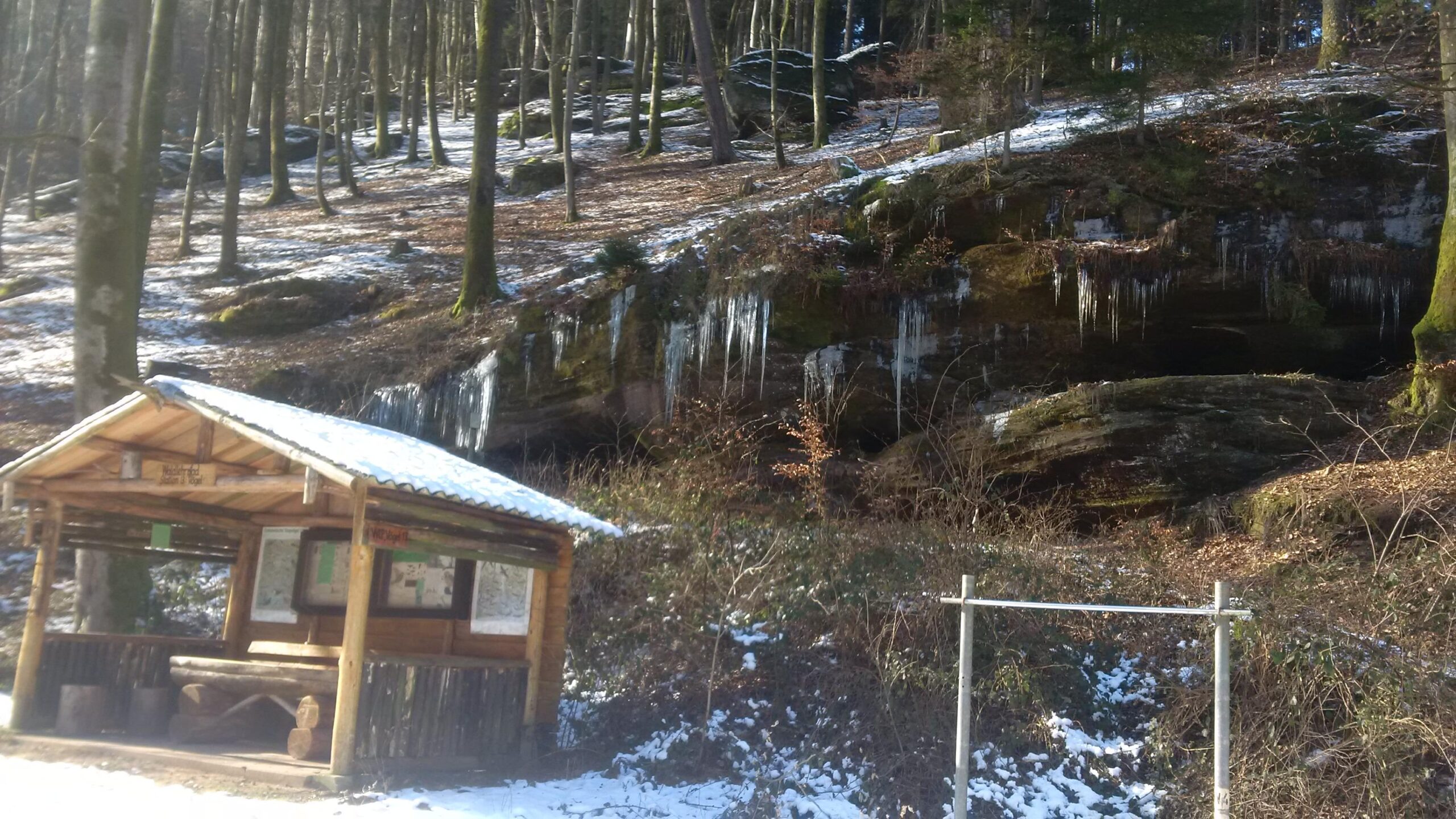 A wooden shelter with a thatched roof is situated in a snowy forest, surrounded by tall trees. In the background, a rock face is partially covered in icicles, and patches of snow lie scattered on the ground. The scene captures a tranquil winter landscape. Countess Sonja Bernadotte Way mountain bike trail.