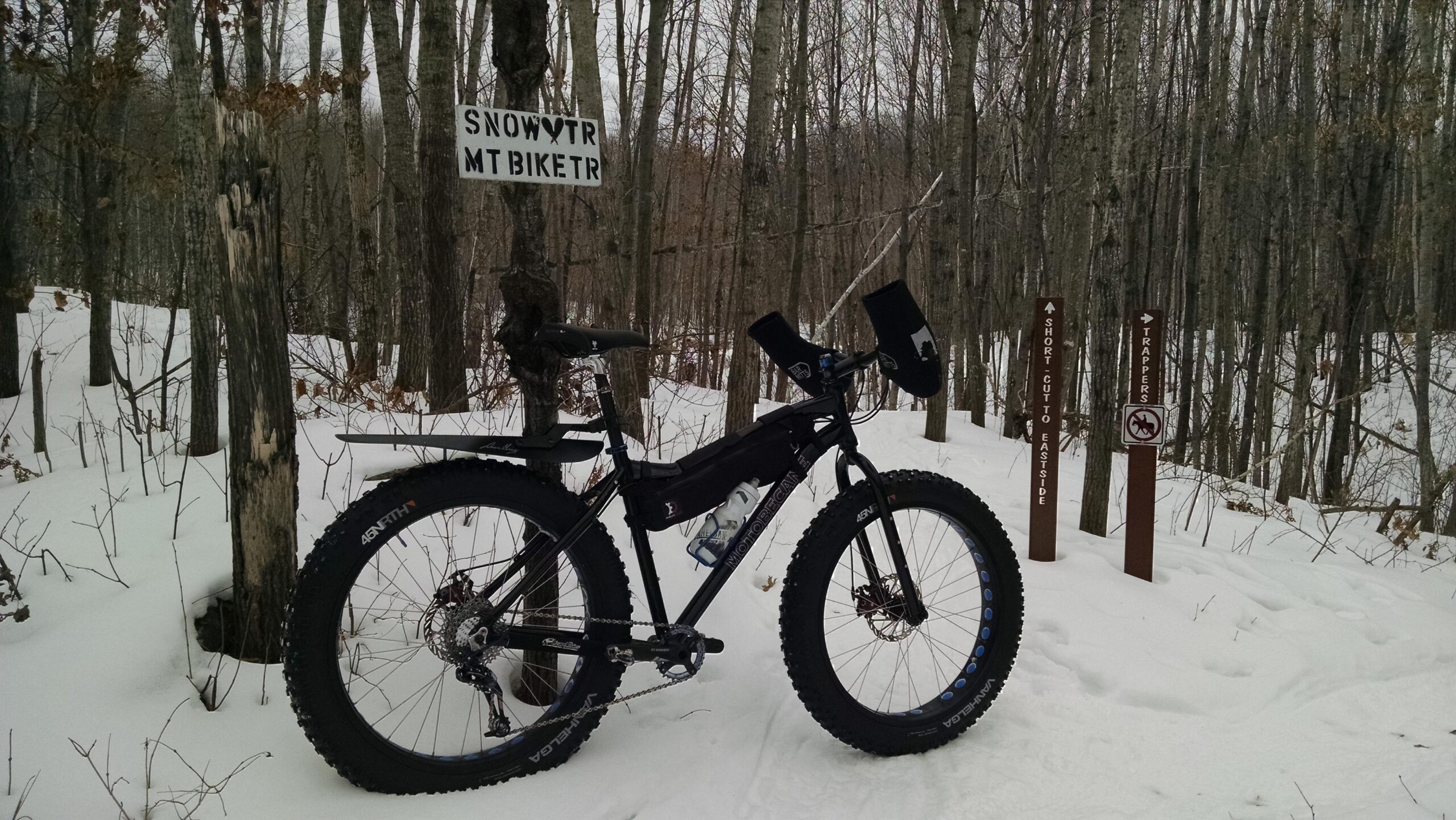 A fat tire mountain bike parked on a snowy trail in a forest. A sign labeled "SNOWTR MT BIKE TR" is visible on a tree, and there are additional directional signs indicating "SHORT CUT TO EASTSIDE" and "TRAPPERS." The surrounding area features a mix of bare trees and patches of snow. Hickory Ridge mountain bike trail.