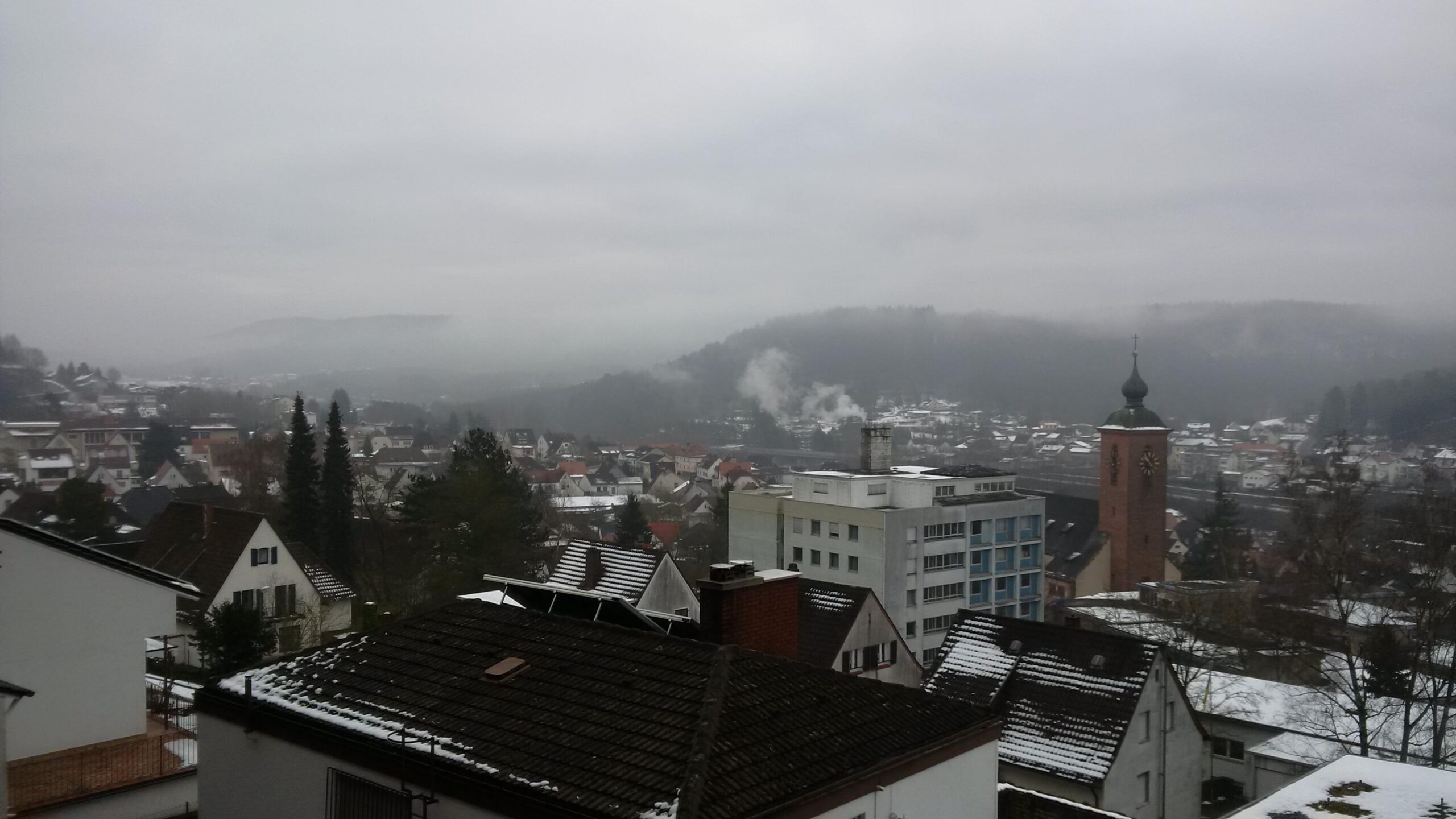 A panoramic view of a small town surrounded by hills on a foggy day. The foreground features rooftops of various buildings, some with a light dusting of snow. A church tower with a clock stands out amidst the houses, while smoke rises from a chimney in the background, blending into the misty atmosphere. The overall scene conveys a quiet, serene ambiance typical of a winter morning. Rodalben F Trail mountain bike trail.