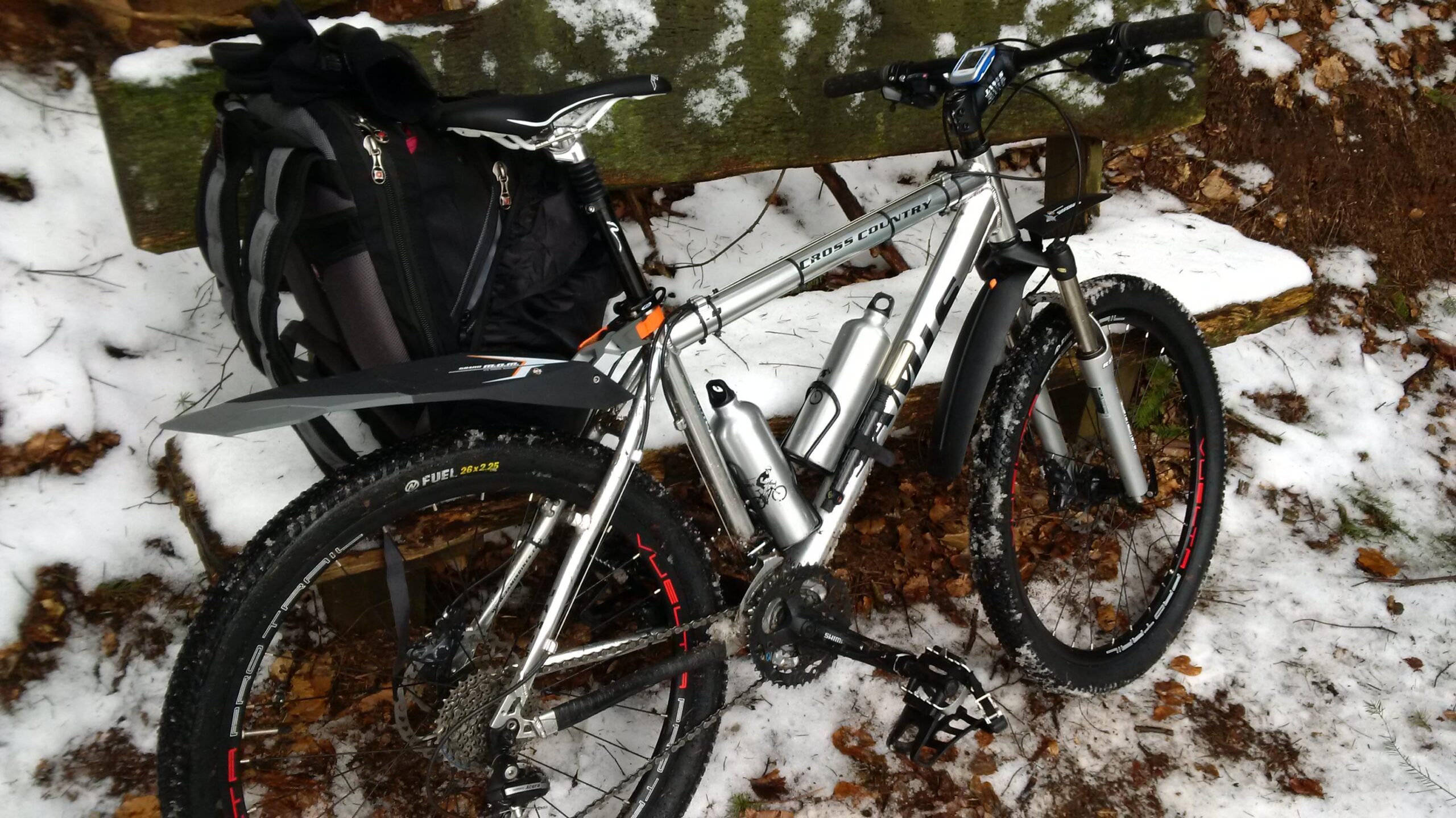 A mountain bike resting against a wooden bench in a snowy setting. The bike features a silver frame, two water bottles mounted on the frame, and is equipped with thick tires suitable for off-road conditions. A black backpack is leaning against the bench, and the ground is covered with a mix of snow and fallen leaves. Rodalben F Trail mountain bike trail.