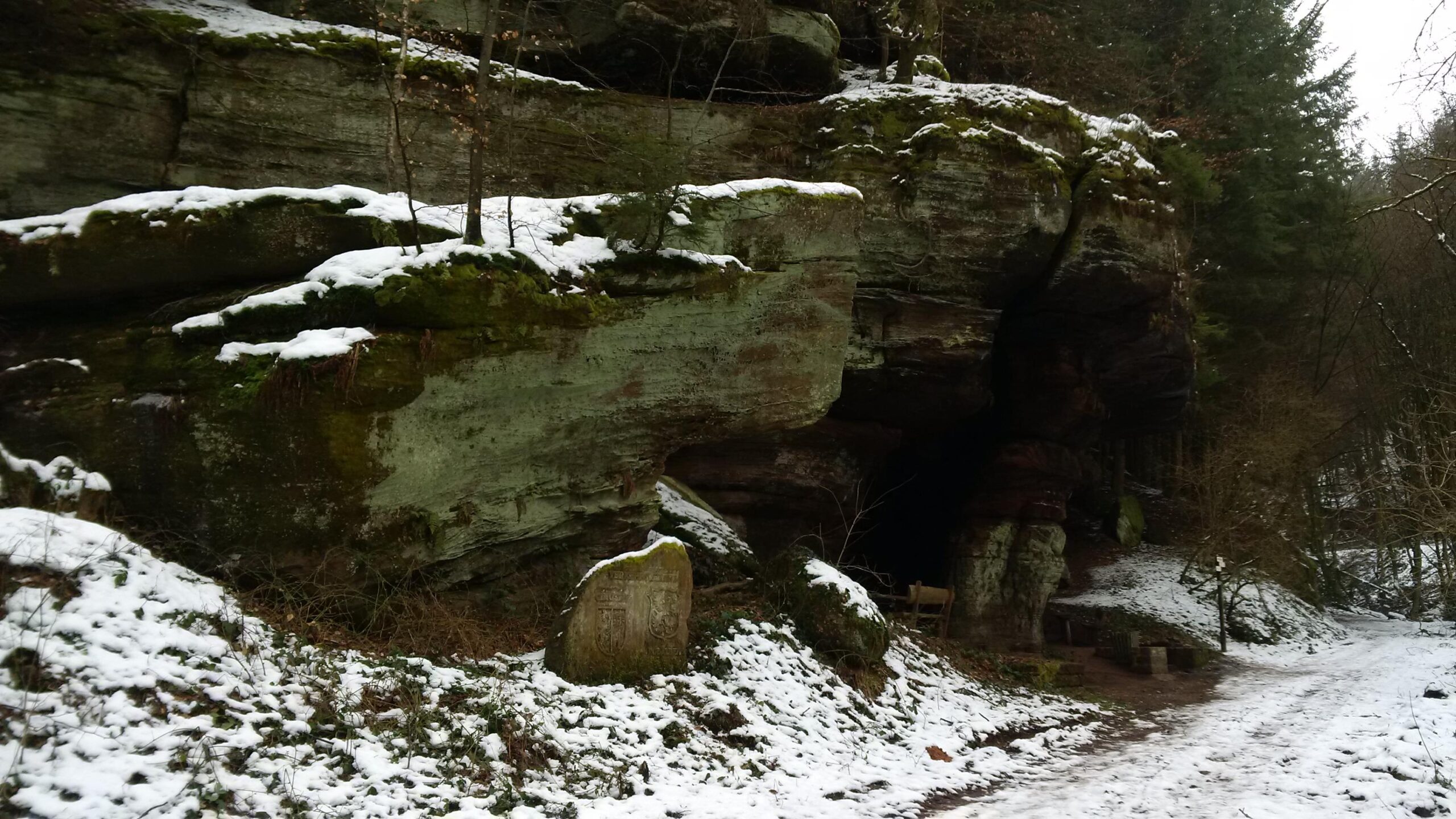 A snow-covered rocky landscape featuring moss-covered boulders and trees. In the foreground, a stone marker is partially visible among the snow, while a winding path leads through the scene. The background is filled with forested hills, creating a serene, wintry atmosphere. Rodalben F Trail mountain bike trail.