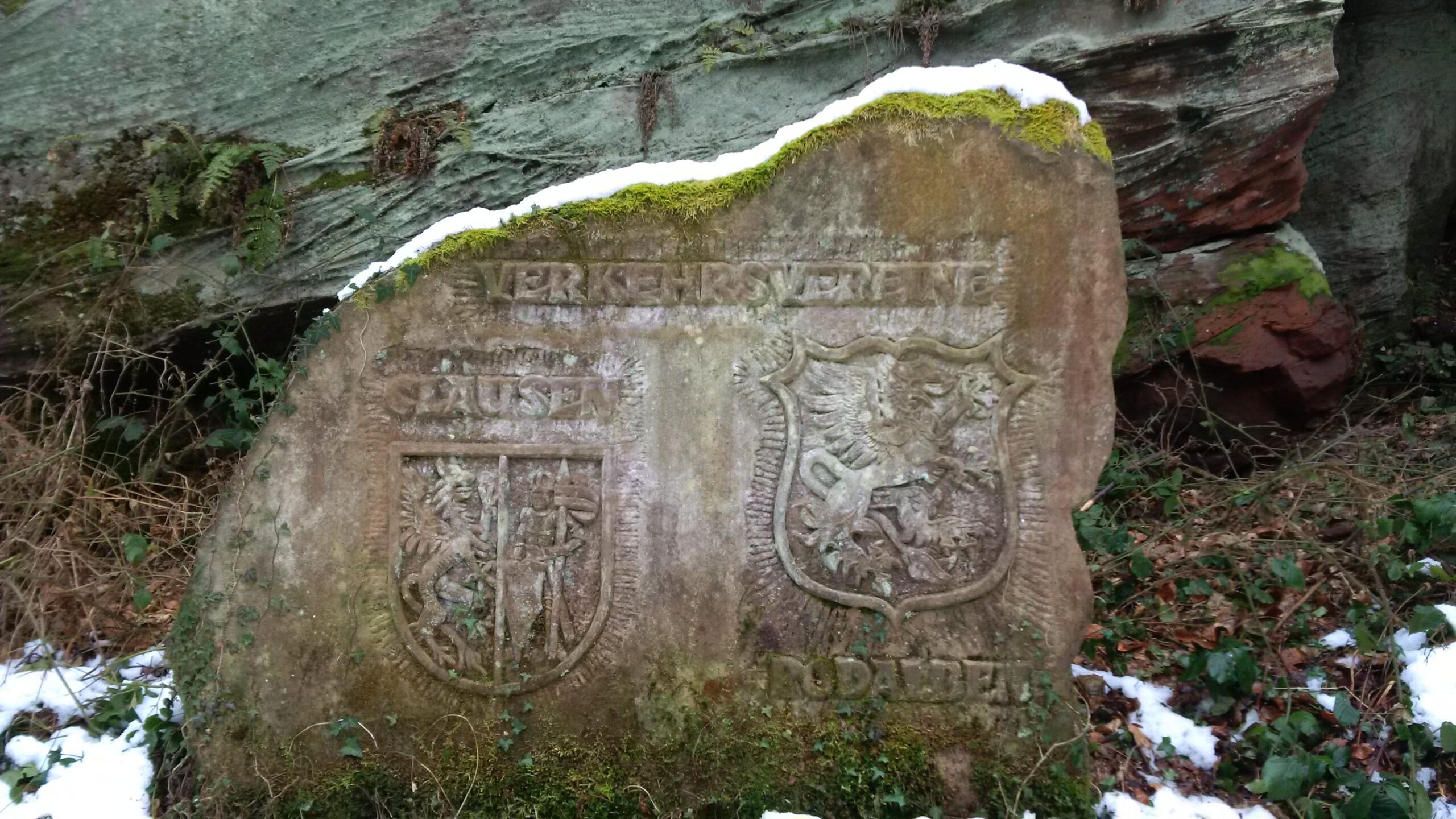 A weathered stone monument featuring two carved coats of arms and inscriptions in German, surrounded by moss, ferns, and a dusting of snow. The stone is situated in a natural setting, partially obscured by greenery and rocks. Rodalben F Trail mountain bike trail.