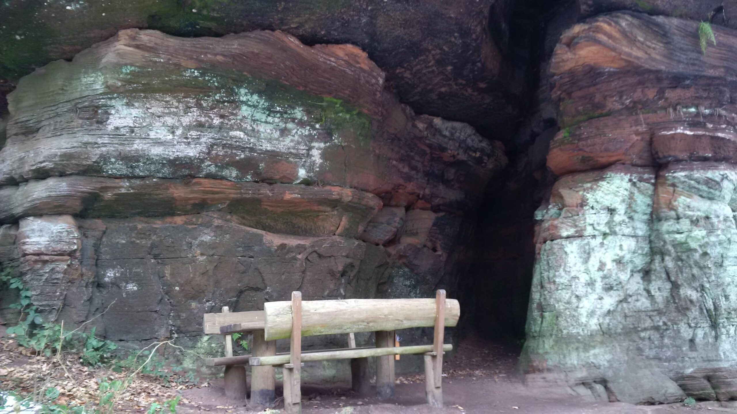 A natural rock formation featuring layered stone with green moss and lichen, alongside a wooden bench in a wooded area. A cave entrance is visible at the back of the formation, surrounded by rugged rocks and foliage. Rodalben F Trail mountain bike trail.