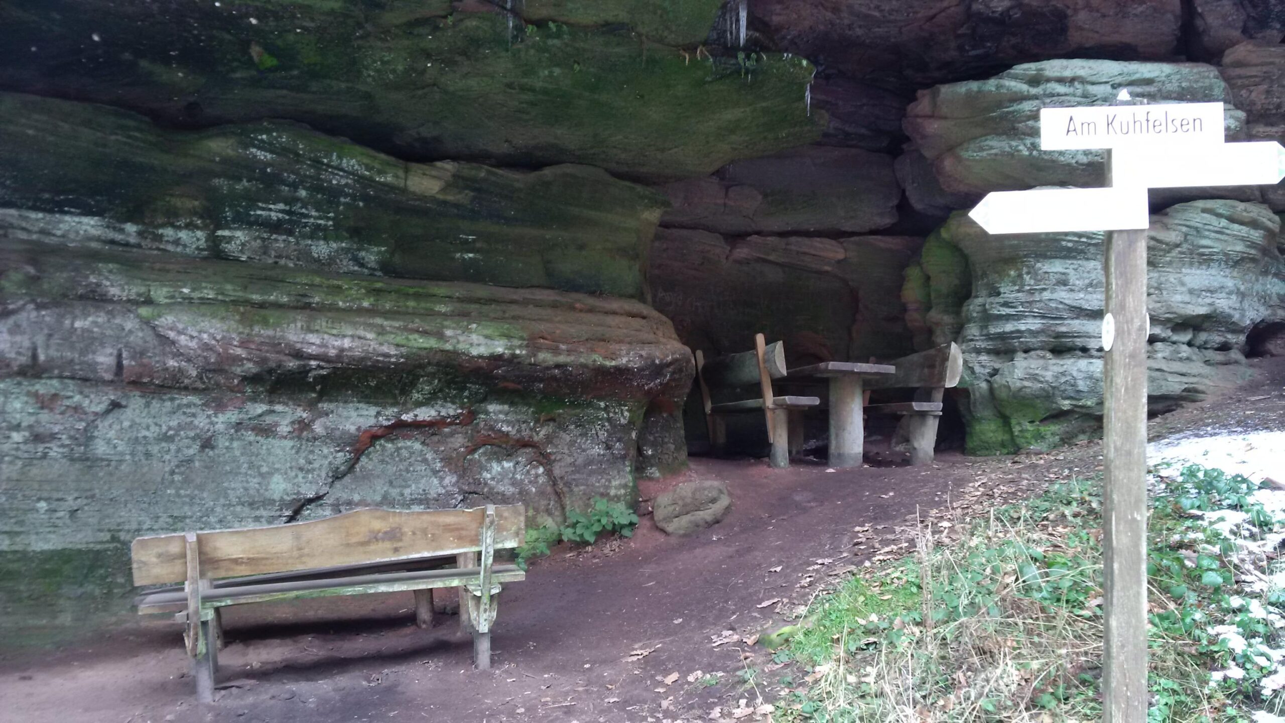 A scenic view of a natural rock formation featuring a shaded seating area with wooden benches and a stone table. A wooden signpost, labeled "Am Kuhfelsen," points in two directions. The surrounding area is lush with green foliage and the ground is covered with fallen leaves, indicating a serene outdoor setting. Rodalben F Trail mountain bike trail.