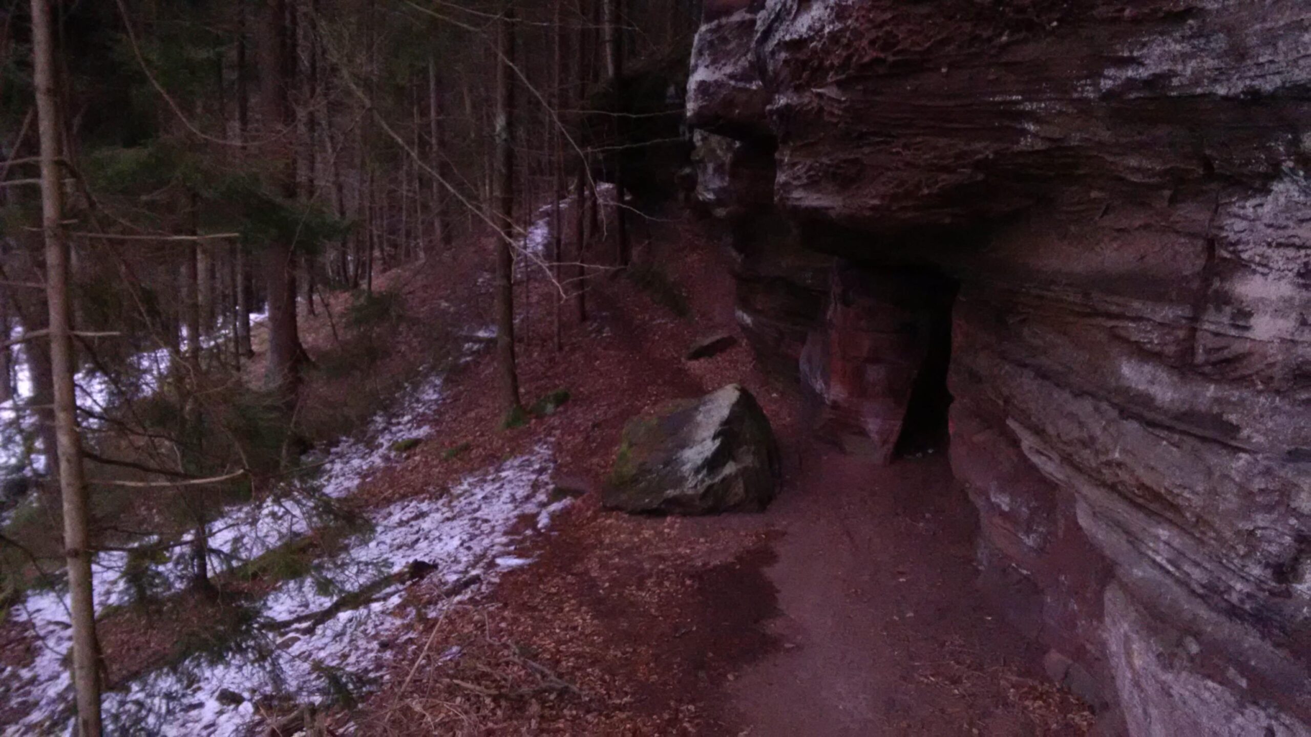 A rocky pathway leading through a forest, with tall trees on either side and patches of snow on the ground. A large boulder is situated on the path, and a cave-like entrance is visible in the rocky outcrop. The scene conveys a tranquil, natural environment. Rodalben F Trail mountain bike trail.