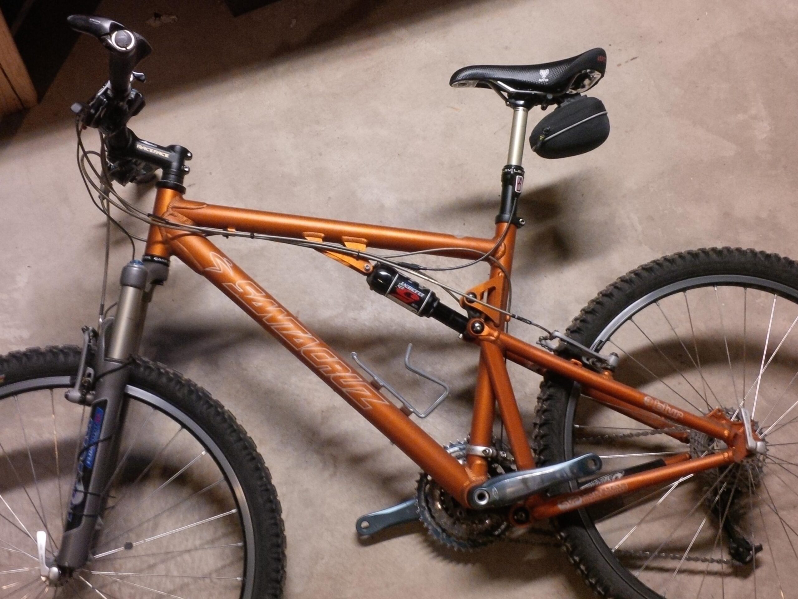 Santa Cruz Blur: A close-up view of a mountain bike with a bright orange frame, featuring a front suspension fork, textured black tires, and a black saddle. The bike is positioned on a concrete floor, highlighting its components such as the handlebars, gear shifters, and several cables.