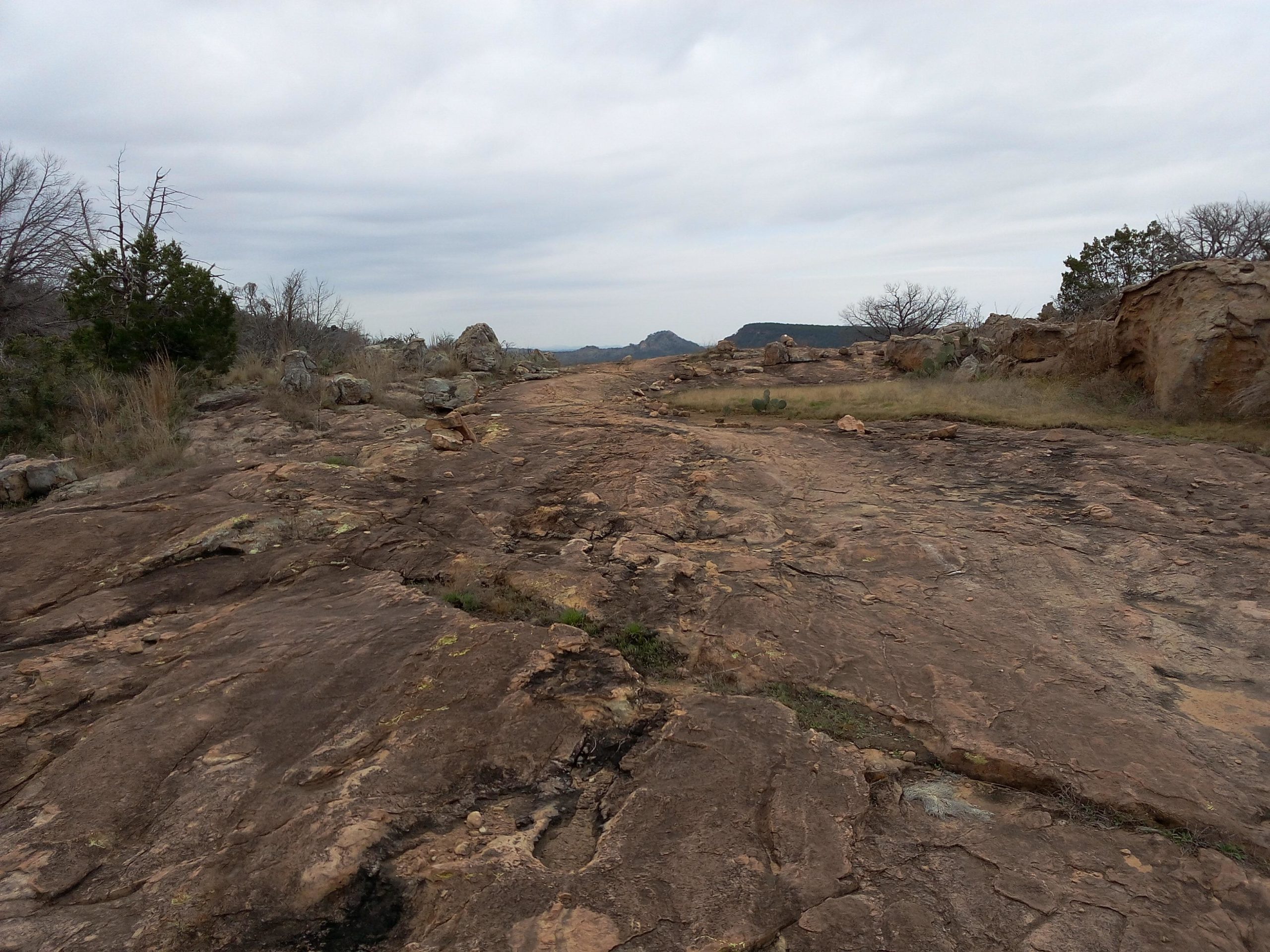 A rocky landscape featuring a broad, flat expanse of weathered stone, with sparse vegetation including a mix of grass and small bushes. In the background, low hills are visible under a cloudy sky, suggesting an overcast day. The scene conveys a natural outdoor setting with rugged terrain. Reveille Peak Ranch mountain bike trail.