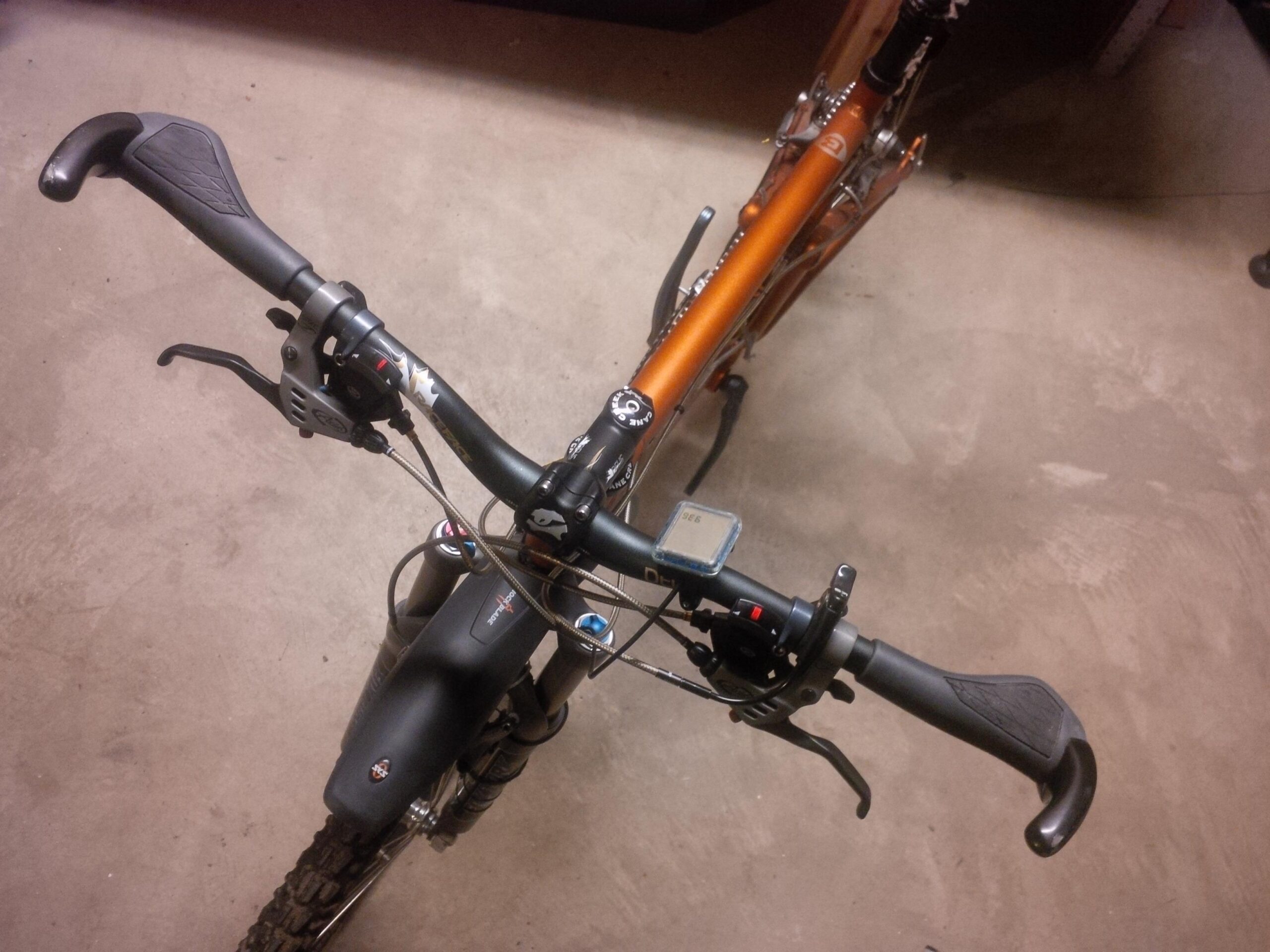 Santa Cruz Blur: A top-down view of a mountain bike's handlebars, showcasing the grips, brake levers, and gear shifters. The bike has a black front fork and a visible speedometer, with parts of the orange frame appearing in the background. The surface beneath the bike is a textured concrete floor.