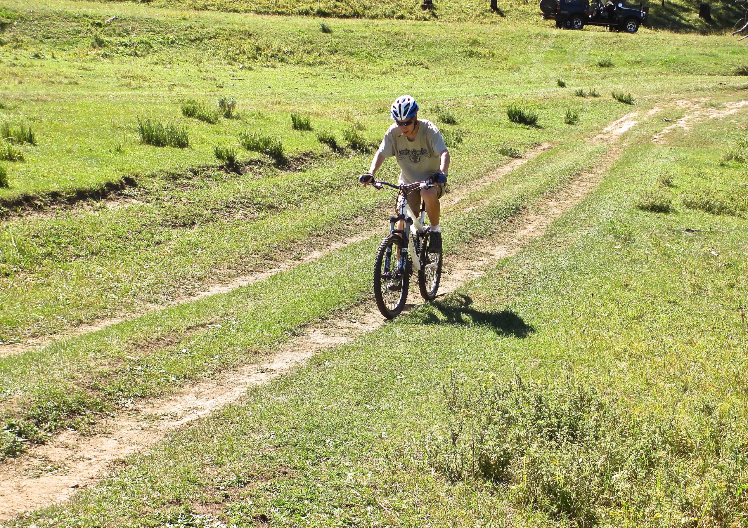 A person riding a mountain bike on a dirt trail surrounded by lush green grass and trees on a sunny day. Khargana Gol mountain bike trail.