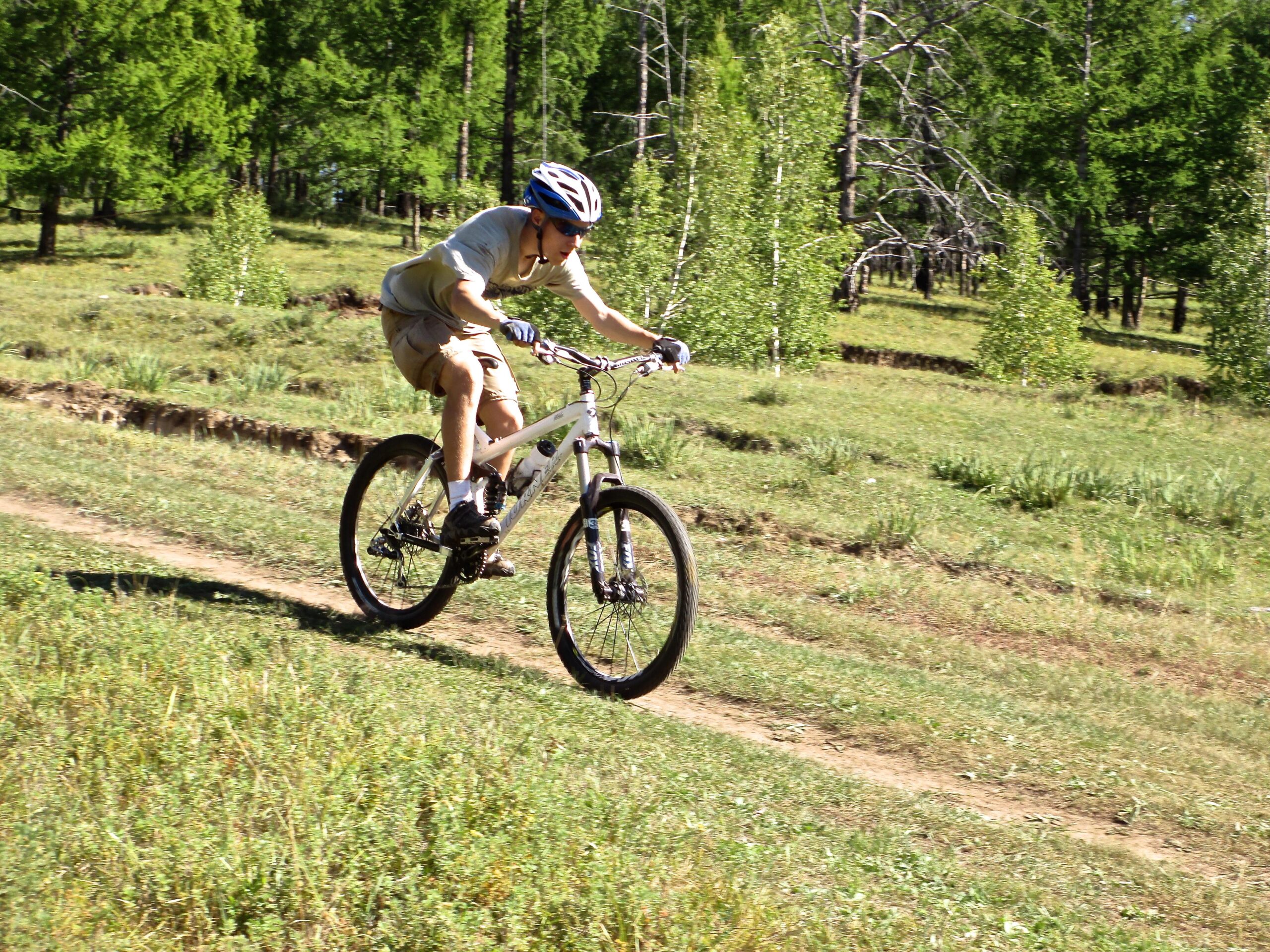 A person riding a mountain bike on a dirt trail through a grassy landscape, surrounded by green trees and shrubs. The cyclist is wearing a helmet and has a focused expression as they navigate the terrain. Khargana Gol mountain bike trail.