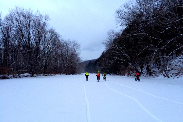 Four cyclists riding fat bikes on a snowy riverbank trail, surrounded by bare trees under a gray sky. The landscape is blanketed in fresh snow, with visible tire tracks leading into the distance.