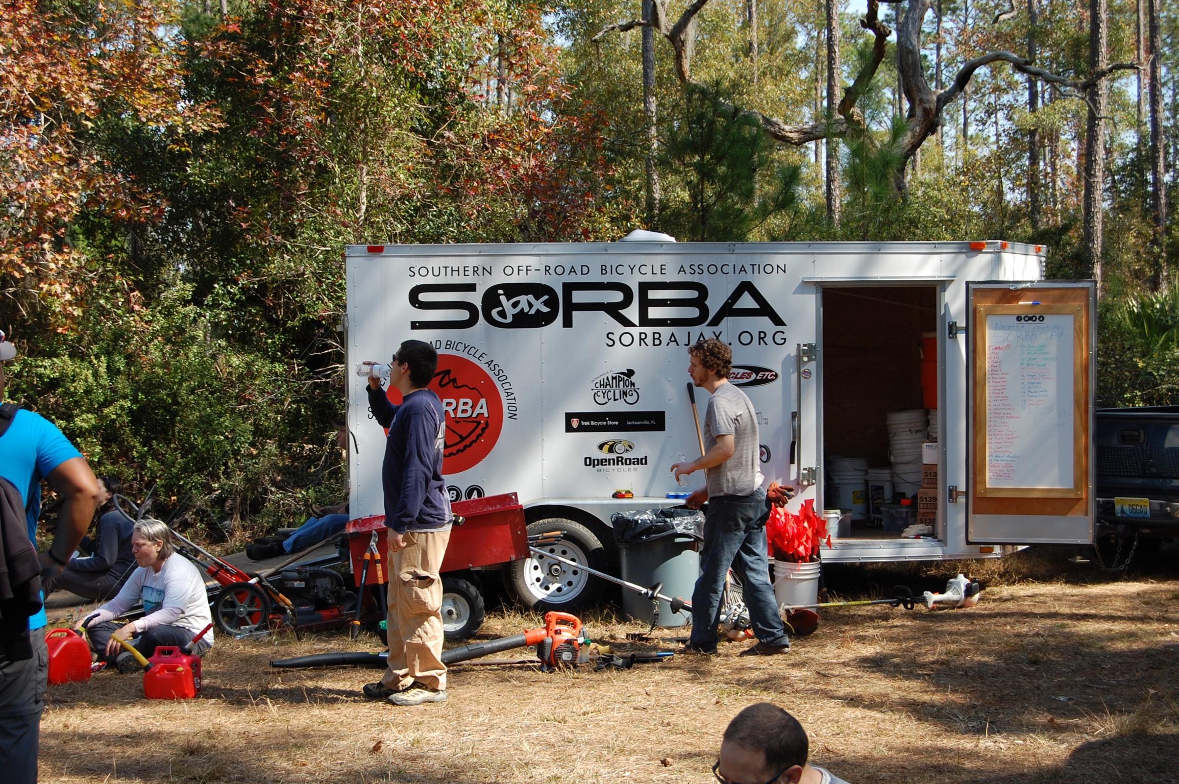 A group of volunteers working on a trail maintenance project, with a trailer displaying the Southern Off-Road Bicycle Association (SORBA) logo in the background. Some individuals are interacting with tools and equipment, while others are resting. The scene is set in a wooded area with autumn foliage. Nocatee mountain bike trail.