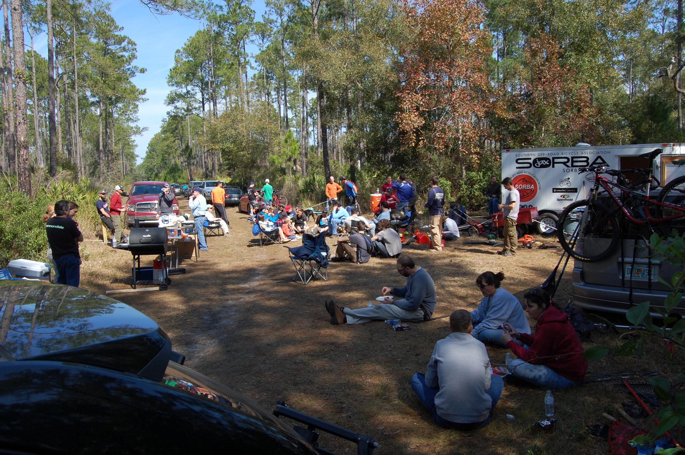 A gathering of people enjoying an outdoor event in a wooded area, with vehicles parked along a dirt path. Participants are engaged in various activities, some seated in lawn chairs while others stand around a grill and enjoy food. A trailer belonging to the Southern Off-Road Bicycle Association (SORBA) is visible in the background, indicating a community event focused on biking and socializing. Nocatee mountain bike trail.