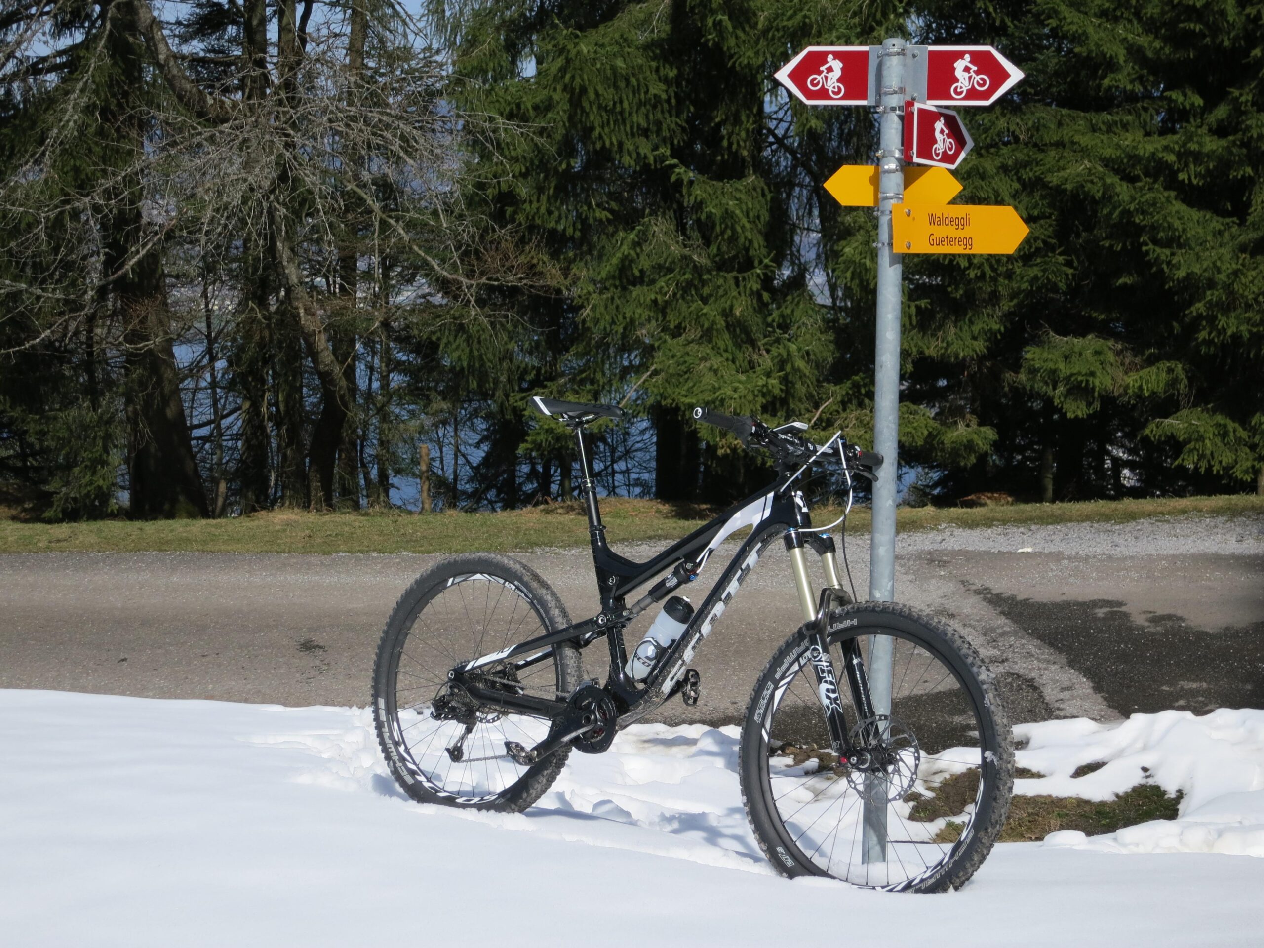 Scott Genius LT 700: A mountain bike leaning against a signpost in a snowy landscape, with various directional arrows indicating cycling paths. In the background, tall trees and a hint of water can be seen, suggesting a scenic outdoor setting.