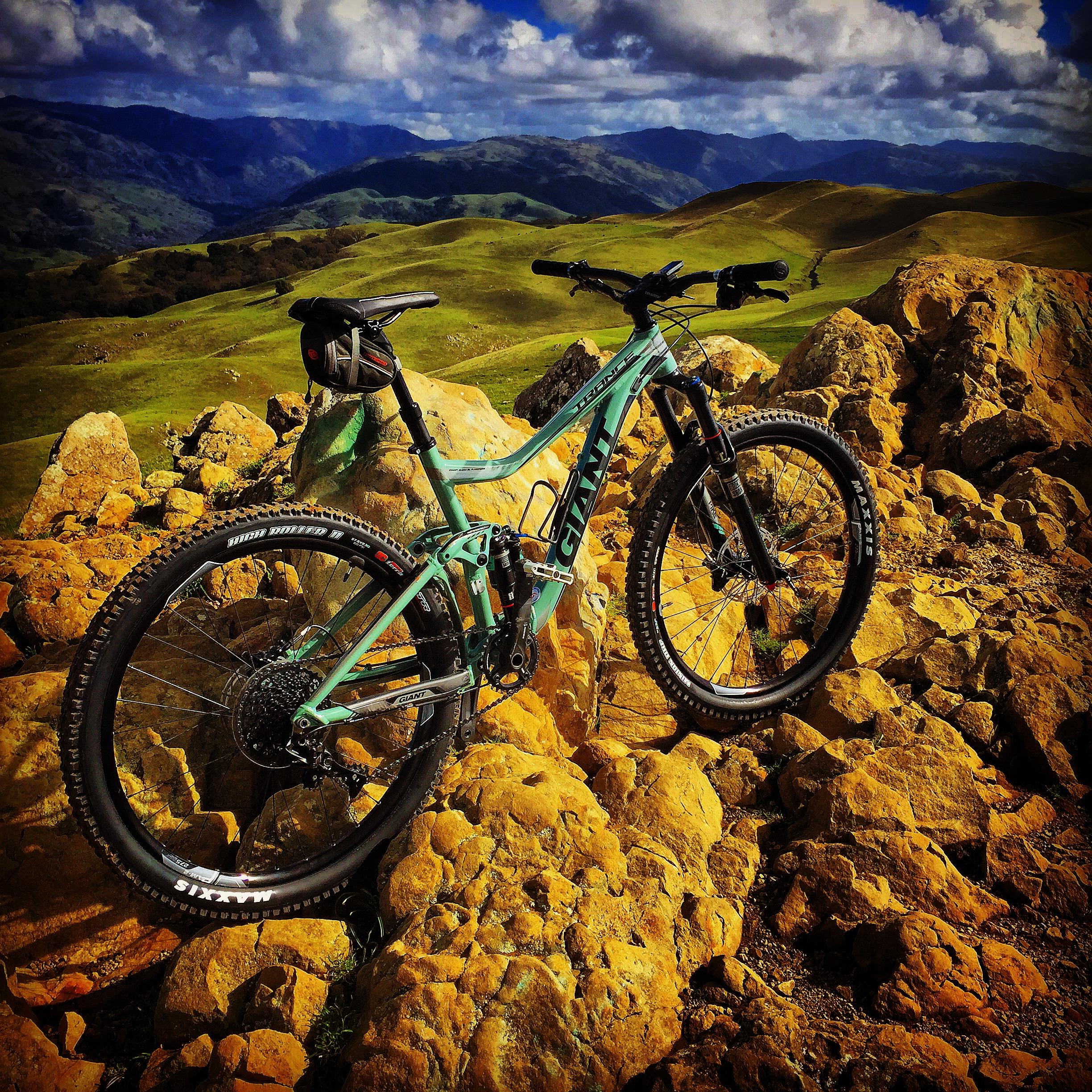 Giant Trance: A turquoise mountain bike parked on rocky terrain, surrounded by lush green hills and a scenic view of distant mountains under a partly cloudy sky.