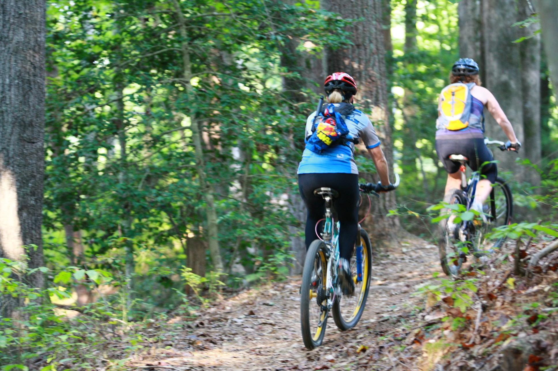 Two cyclists riding on a dirt trail through a forest, surrounded by lush green trees. One cyclist is wearing a blue shirt and a colorful backpack, while the other is in a sleeveless top and black shorts. Sunlight filters through the trees, creating a serene outdoor atmosphere. Overmountain Victory Trail mountain bike trail.