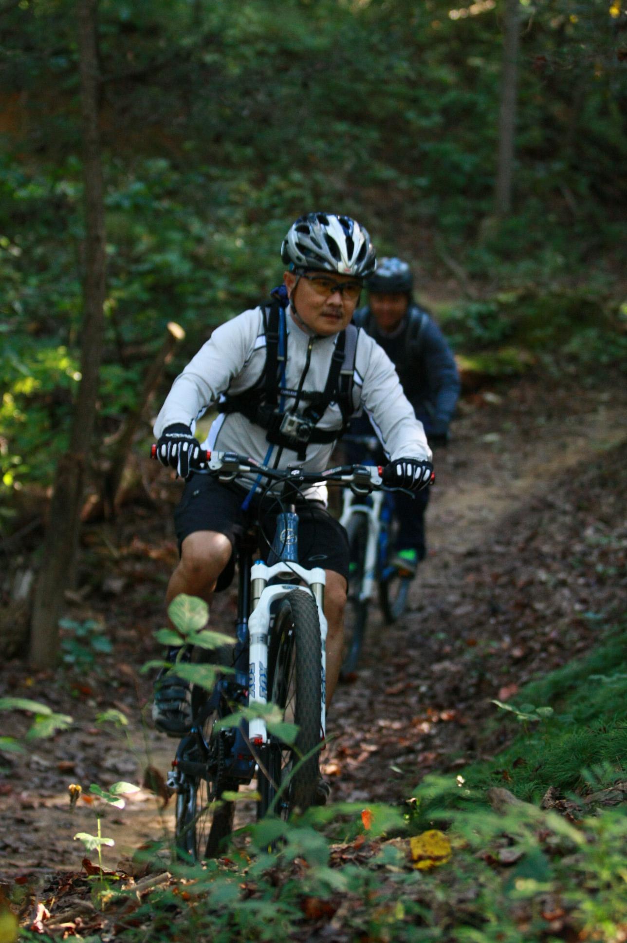 Two mountain bikers riding on a dirt trail surrounded by greenery. One biker, wearing a white long-sleeve shirt and a helmet, is in the foreground, focused on the path ahead. The second biker, in a blue jacket, follows closely behind. The scene captures a vibrant autumn landscape with fallen leaves on the ground. Overmountain Victory Trail mountain bike trail.